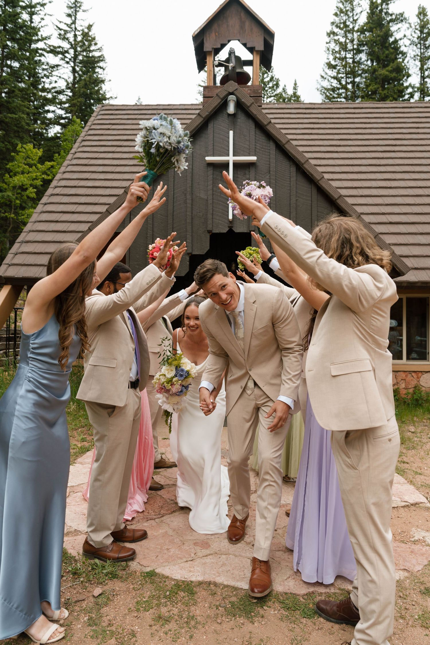 Wedding party celebrating outside rustic mountain chapel with raised bouquets.