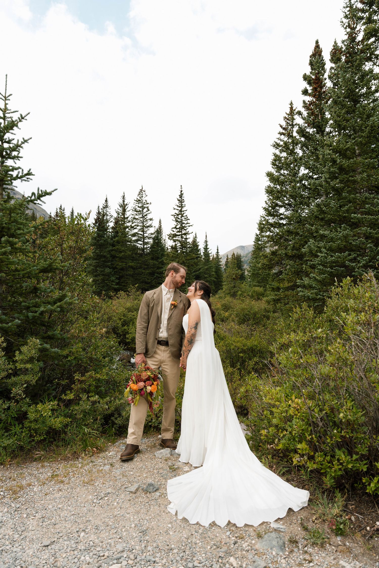 Bride and groom share a romantic moment on a mountain trail surrounded by evergreen trees in a stunning outdoor wedding.