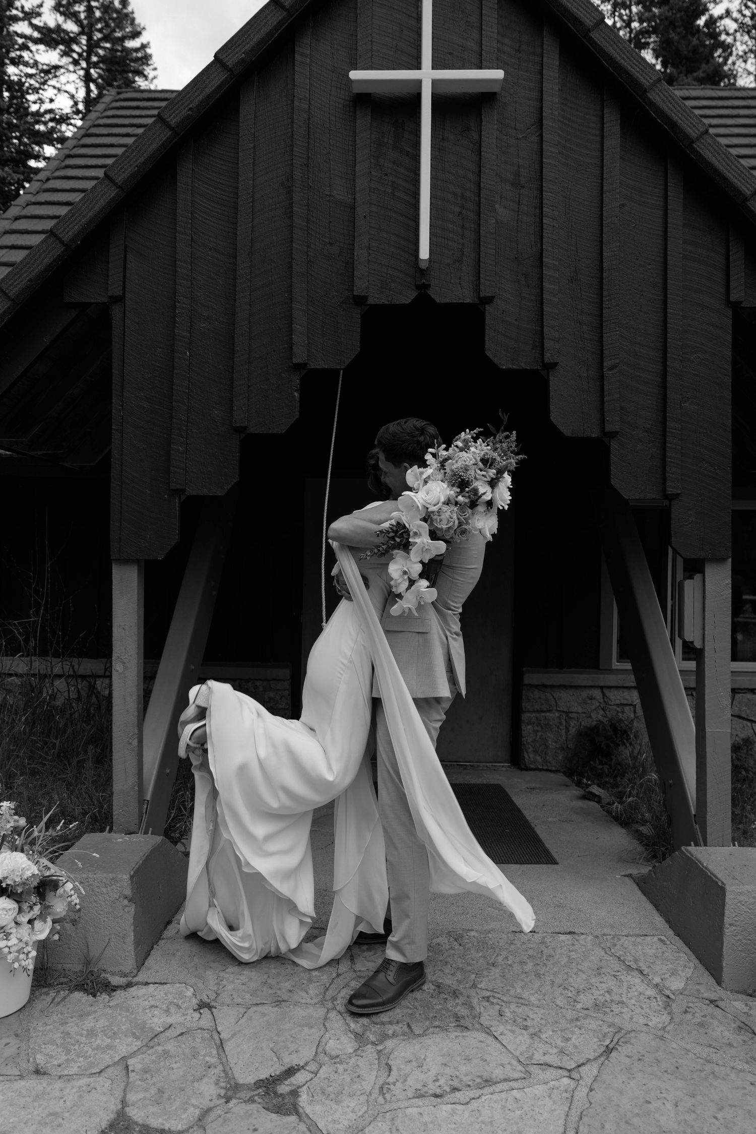 Black and white photo of bride and groom kissing outside a rustic chapel with a cross, bride holding a floral bouquet.