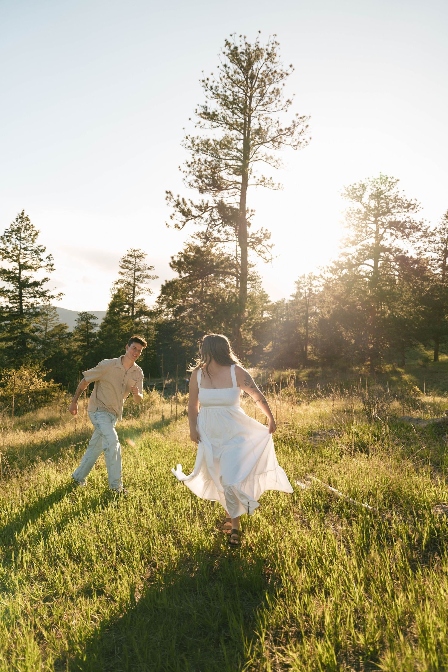 Couple running through sunlit meadow with tall pine trees, woman in white dress, golden hour light.