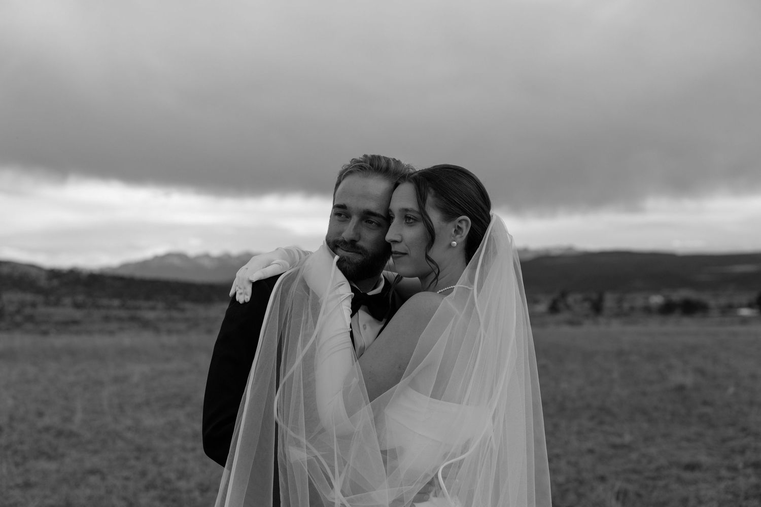 Black and white photo of a bride and groom embracing in an open field with dramatic cloudy skies.
