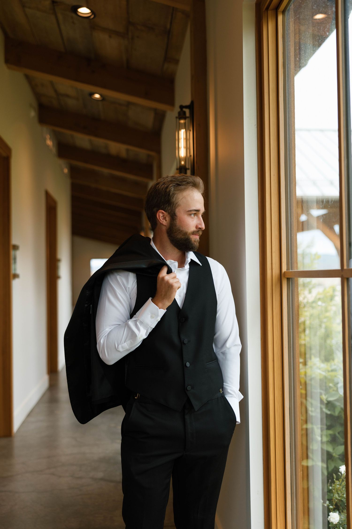 Handsome bearded groom in black vest holding suit jacket while gazing out window.