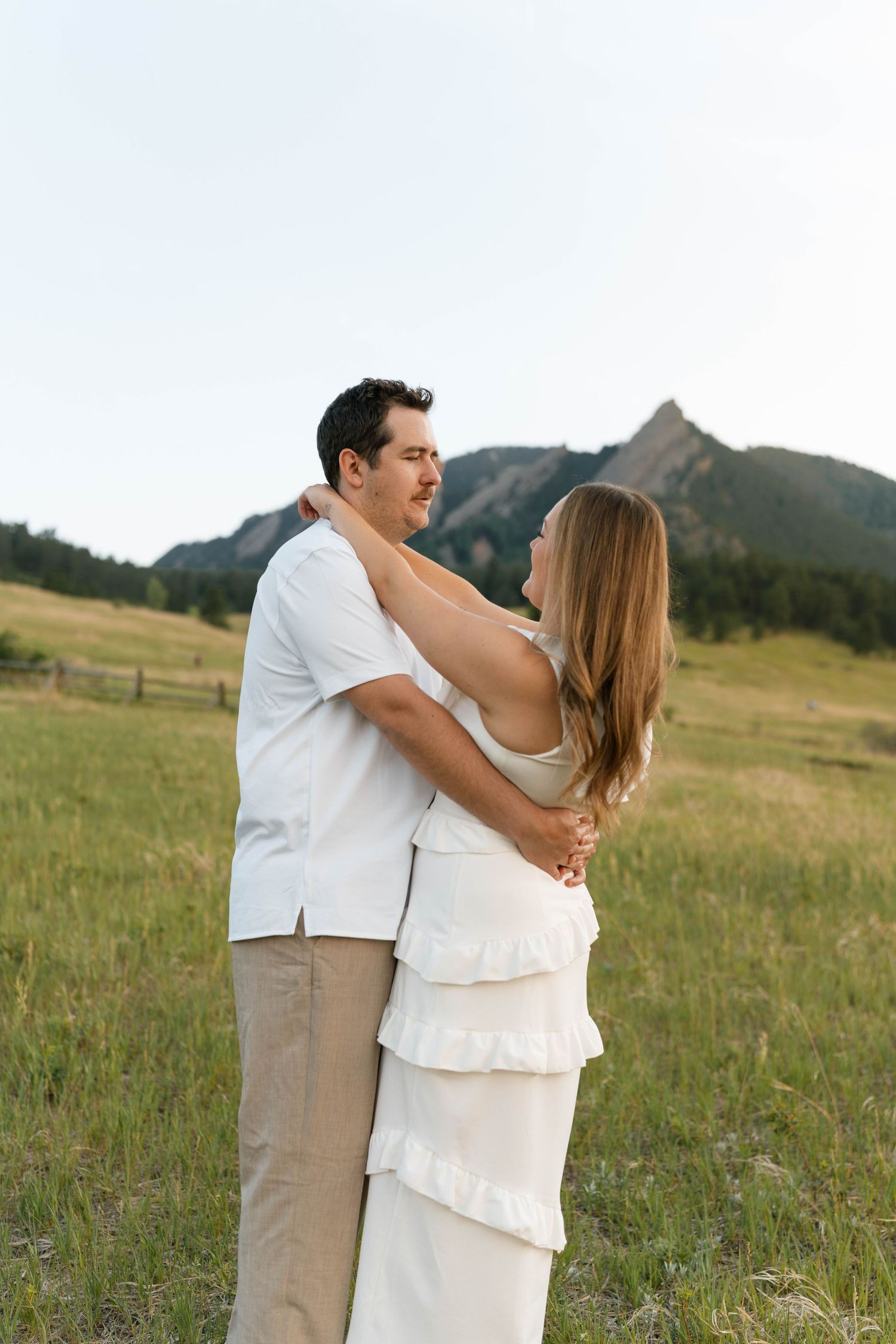 A couple embraces in a green meadow with rocky mountain peaks in the background on a clear day.