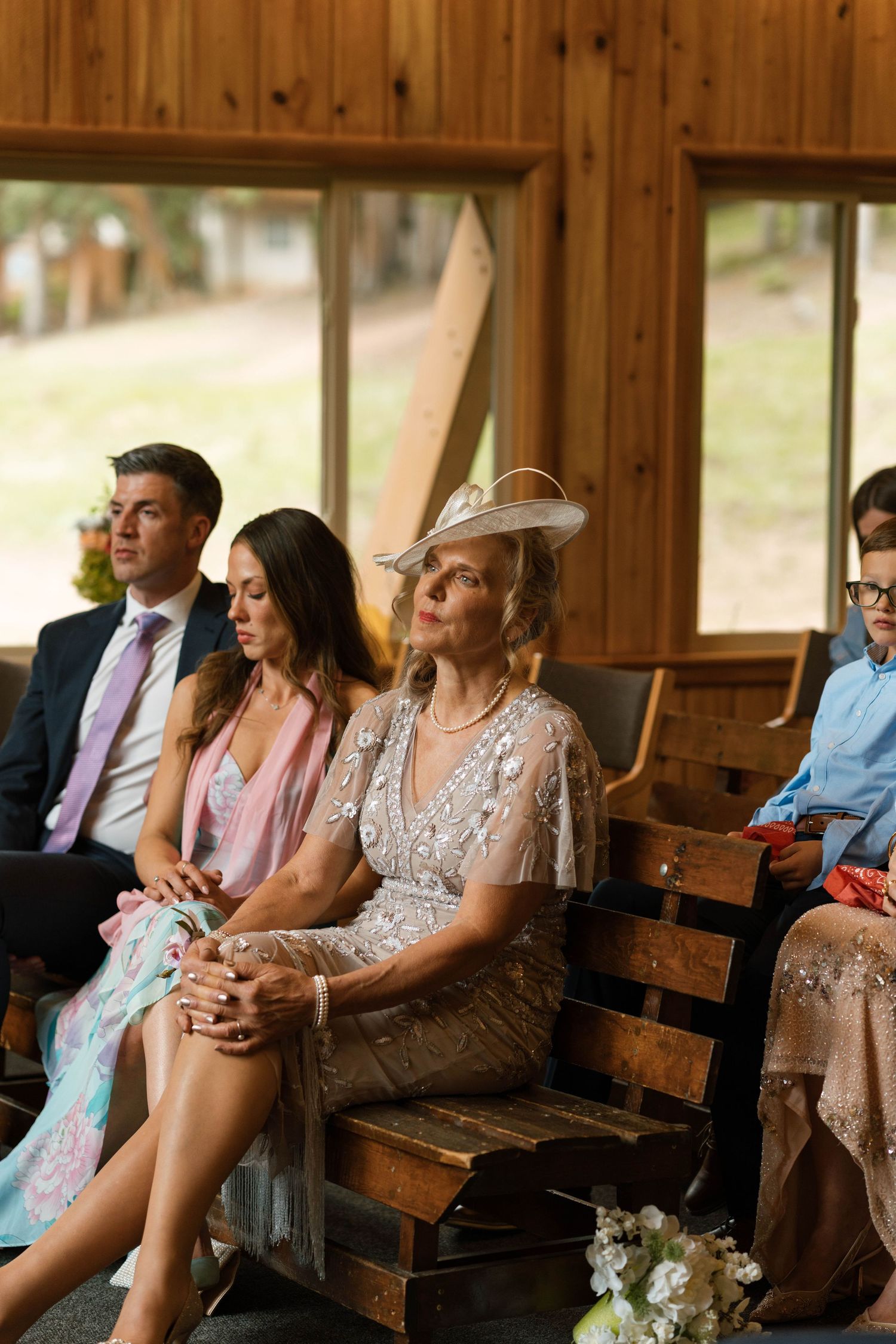 Elegantly dressed wedding guests seated in a wood-paneled chapel during a wedding ceremony.