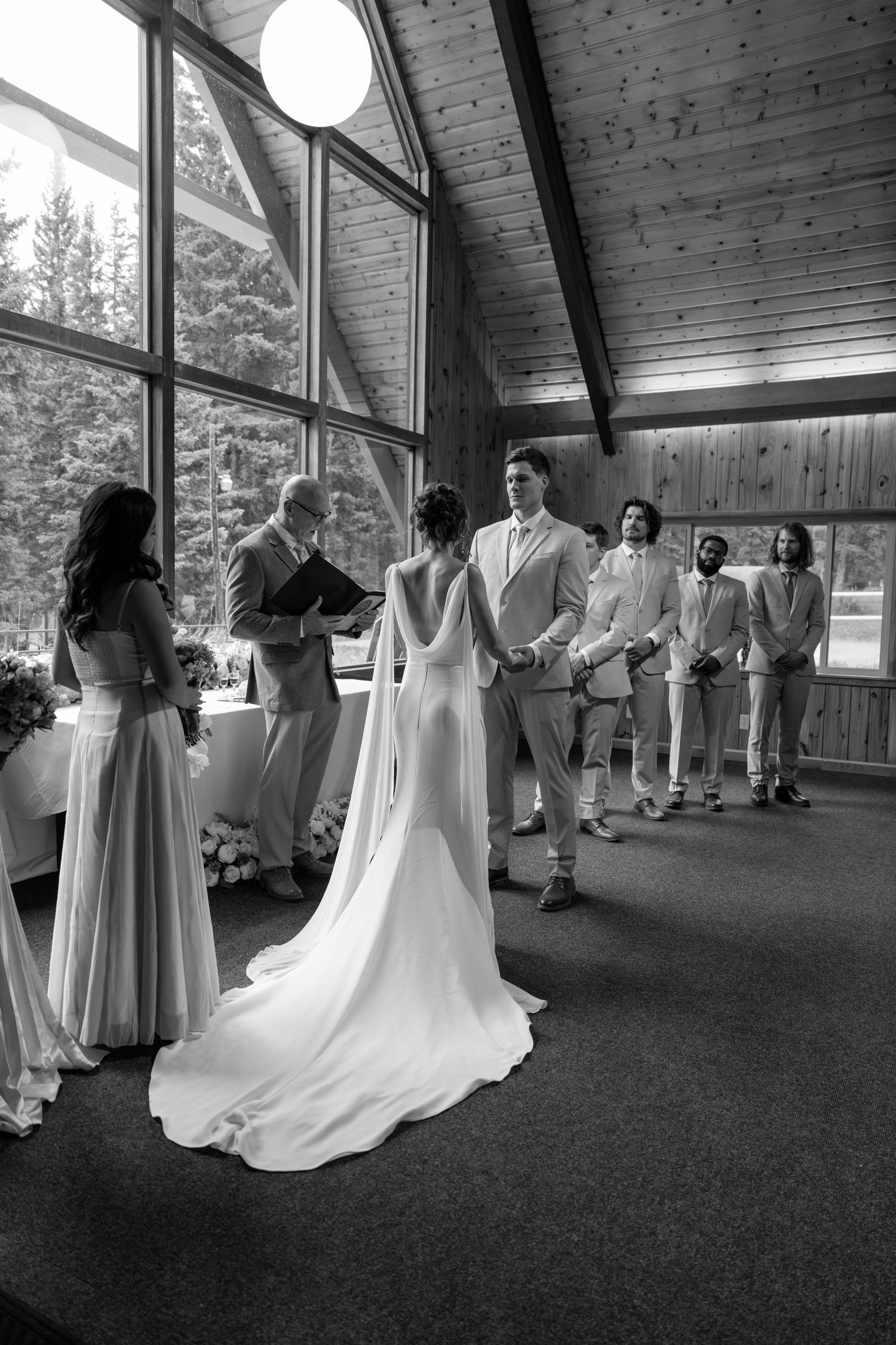 Black and white photo of a bride and groom exchanging vows in a chapel with groomsmen and bridesmaids.