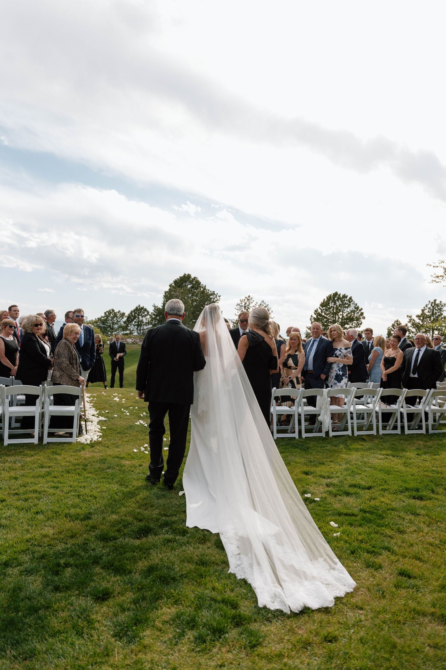 Wedding ceremony on a hilltop lawn with white chairs and flowing bridal veil against cloudy sky.