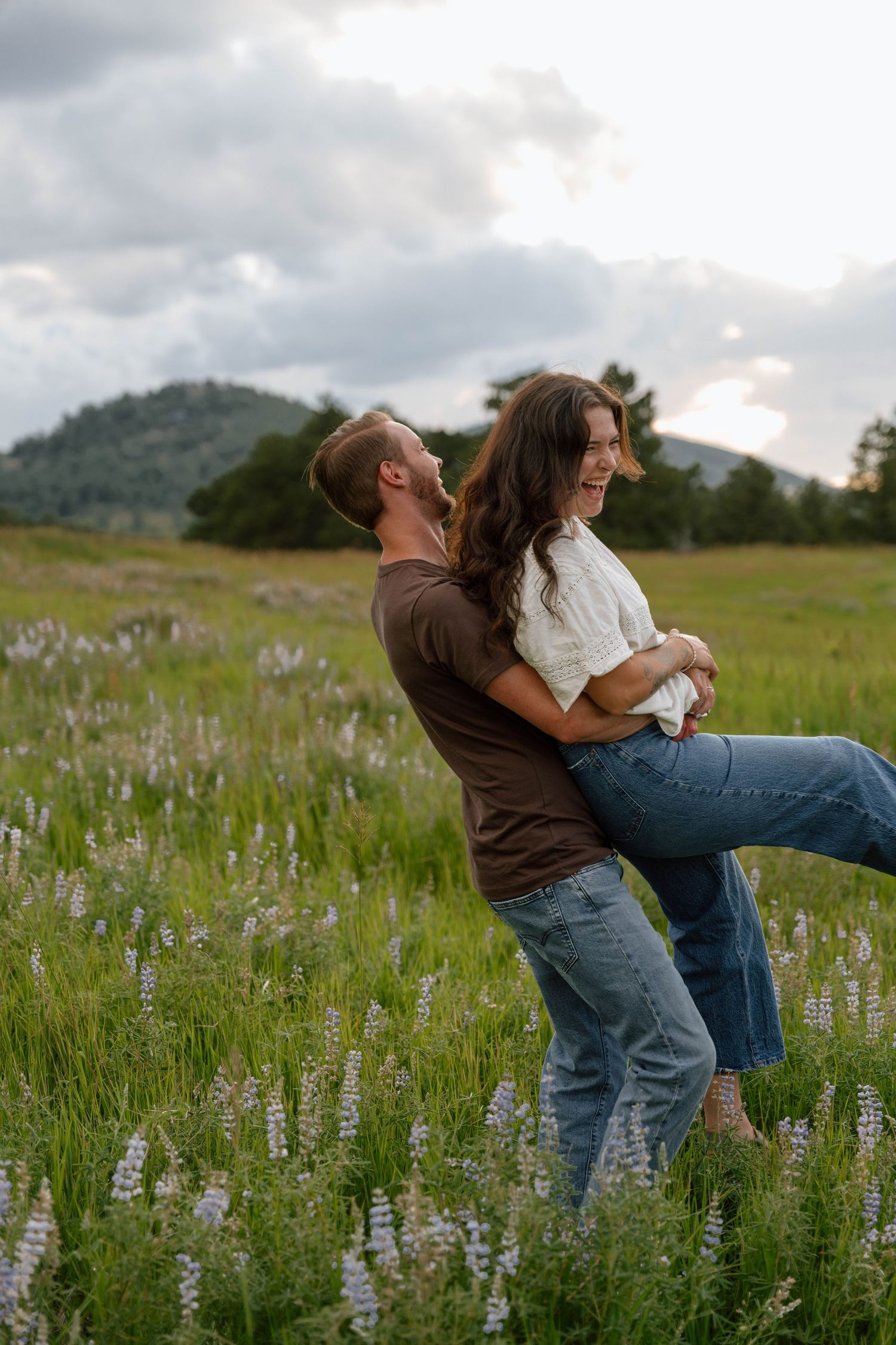 A couple shares playful moments in a wildflower meadow with mountains in the background at sunset.