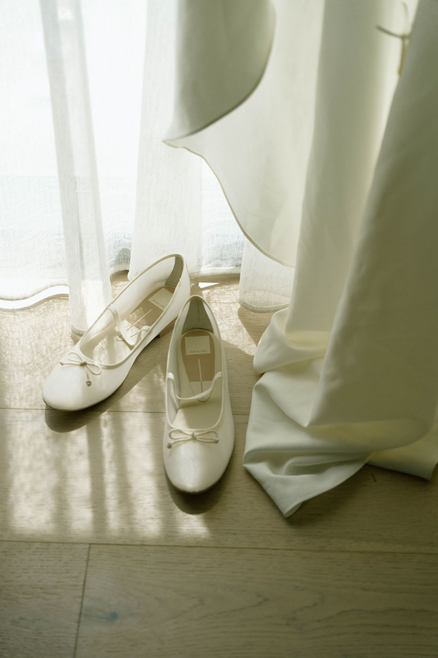 White ballet flats rest on hardwood flooring beside flowing white curtains in natural light.