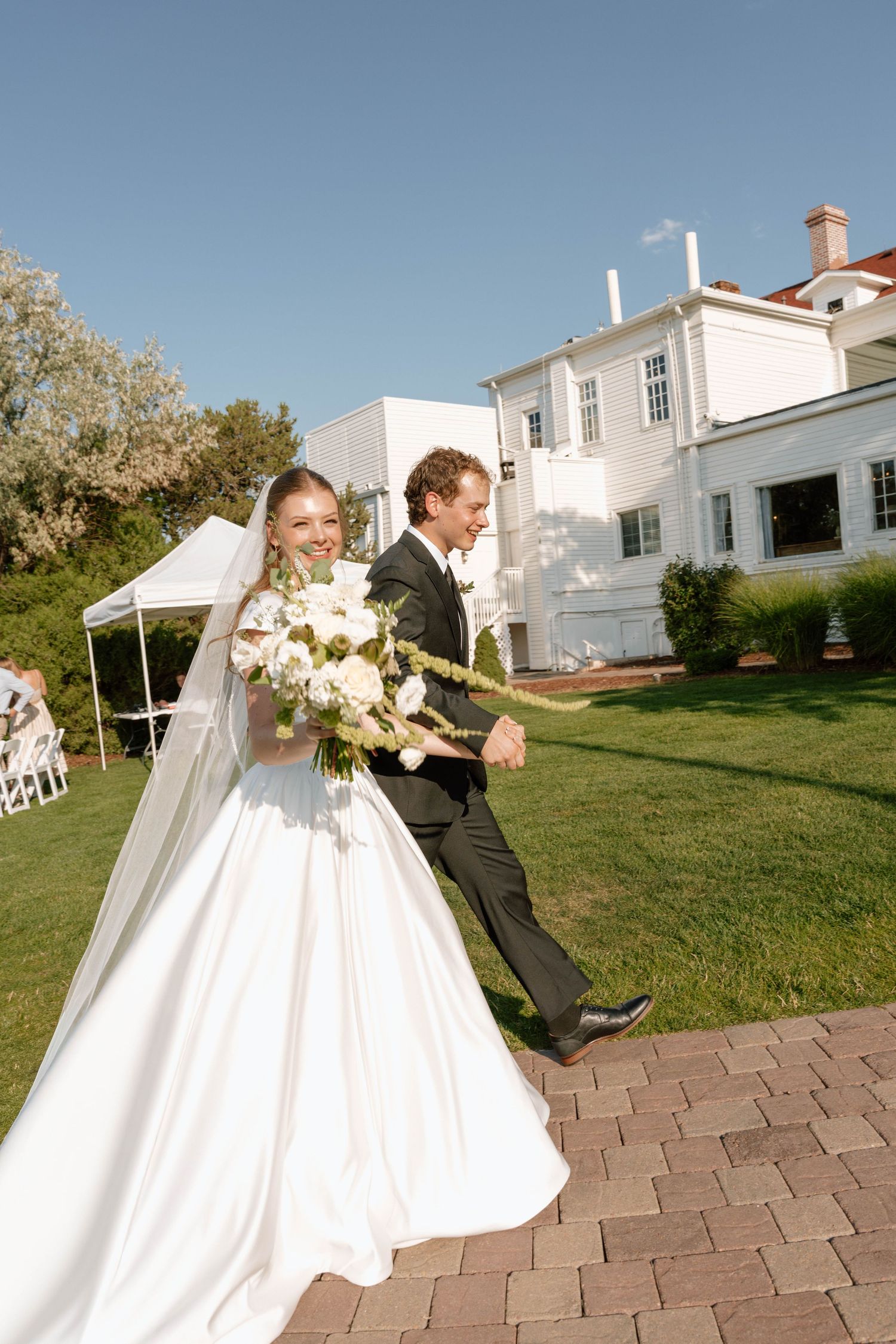 A wedding couple walks together on a brick path in front of a white historic mansion on a sunny day.