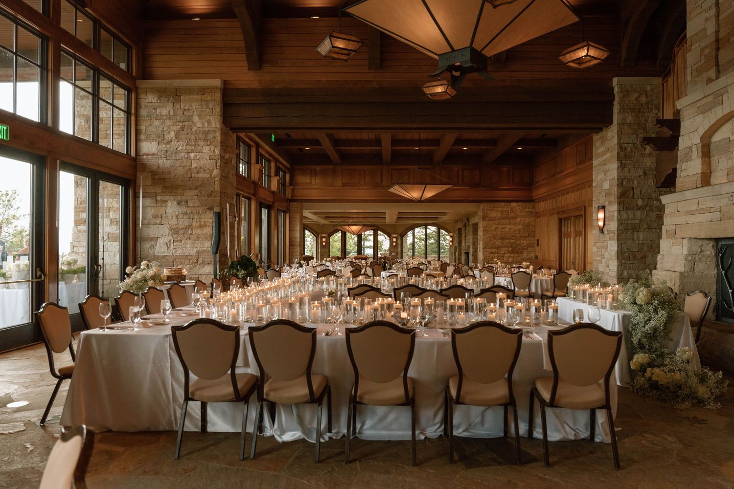 Elegant wedding reception hall with long banquet tables, candles, floral centerpieces, wood beams, and stone walls.