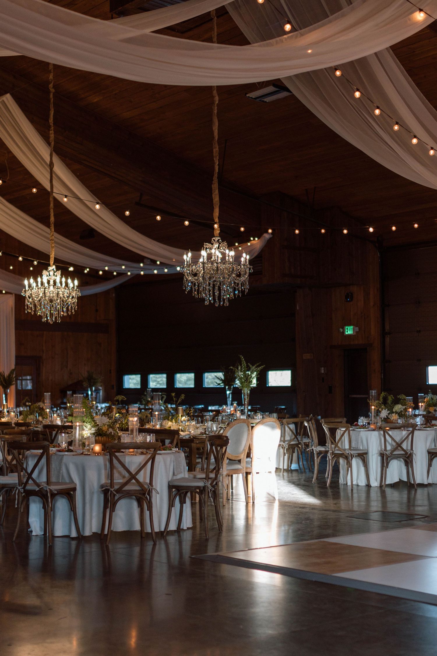 Elegant barn wedding reception with crystal chandeliers, string lights, white draping, round tables, and a wooden dance floor.