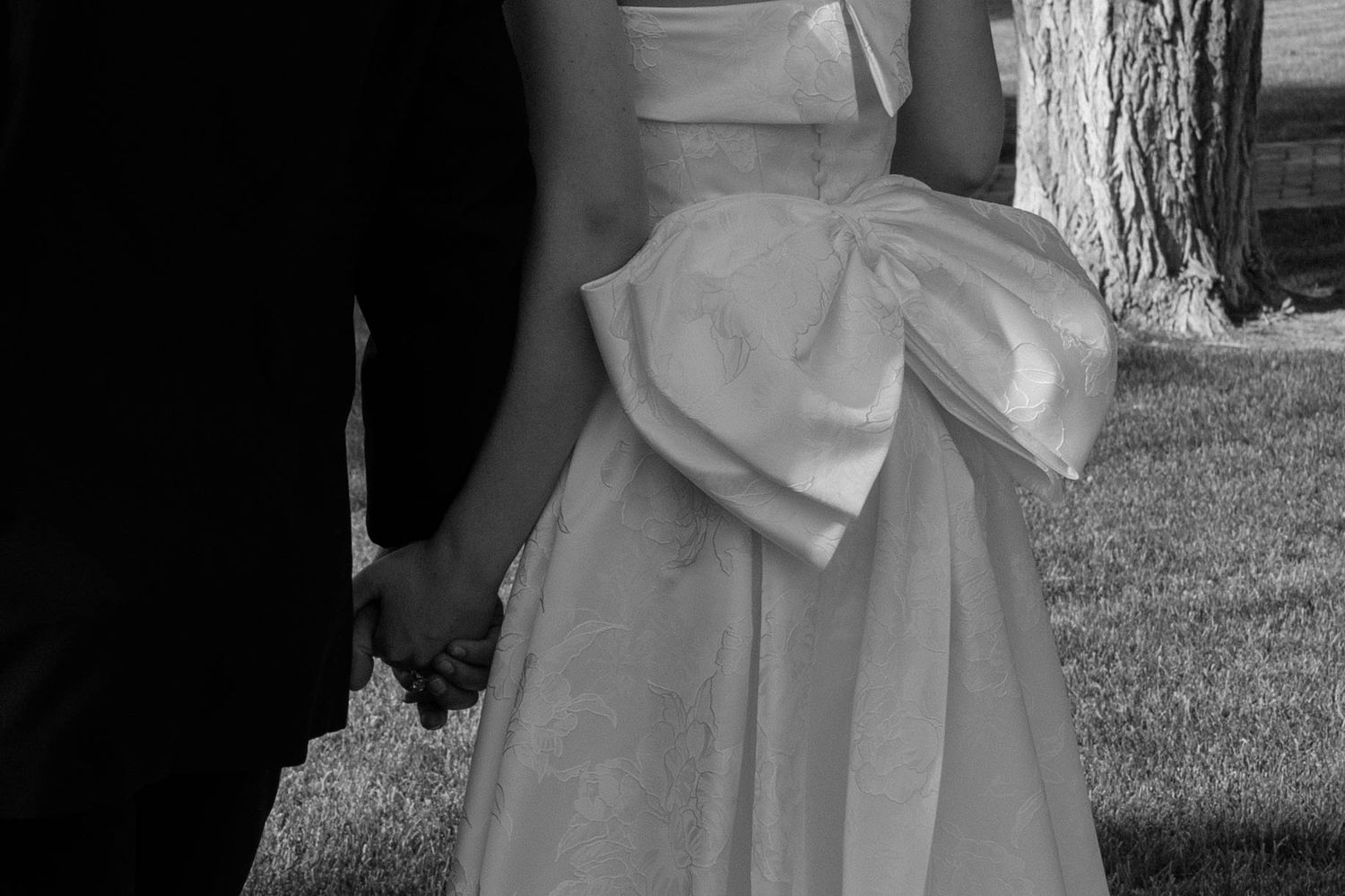 Black and white photo of a bride in a white gown with bow holding hands with groom in dark suit outdoors.