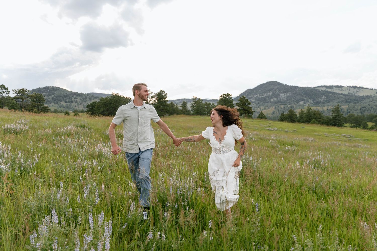 A couple walks hand in hand through a scenic mountain meadow filled with vibrant purple wildflowers on a sunny day.