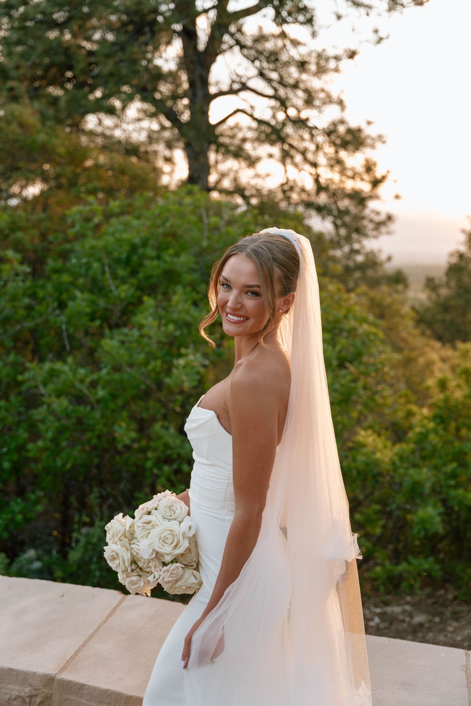 Elegant bridal portrait outdoors with white roses and flowing veil at sunset.