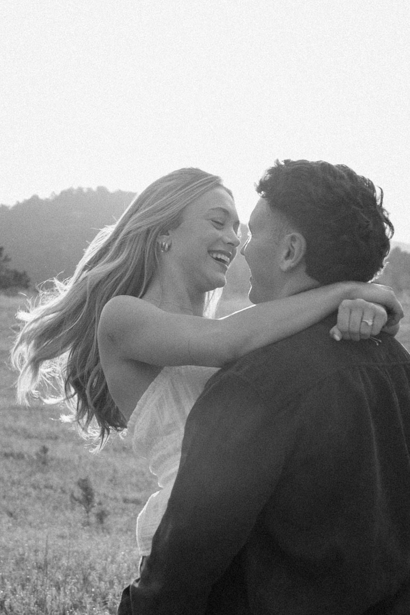 Black and white photo of a joyful couple embracing outdoors, woman laughing with hair blowing in the wind.