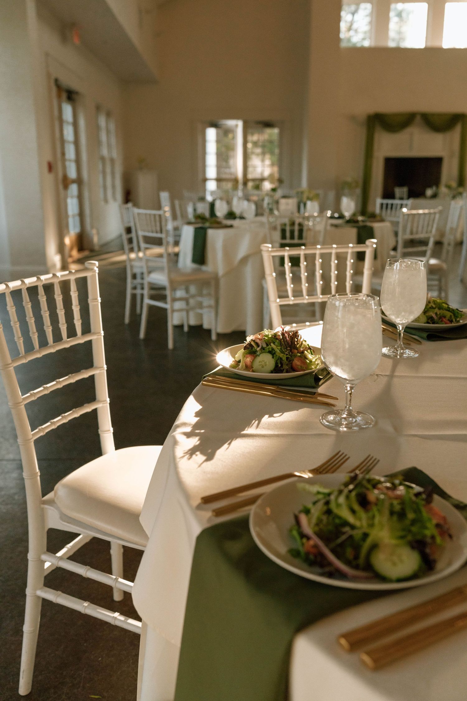 Elegant banquet hall with white Chiavari chairs, green table runners, gold cutlery, and fresh salads set for a formal event.