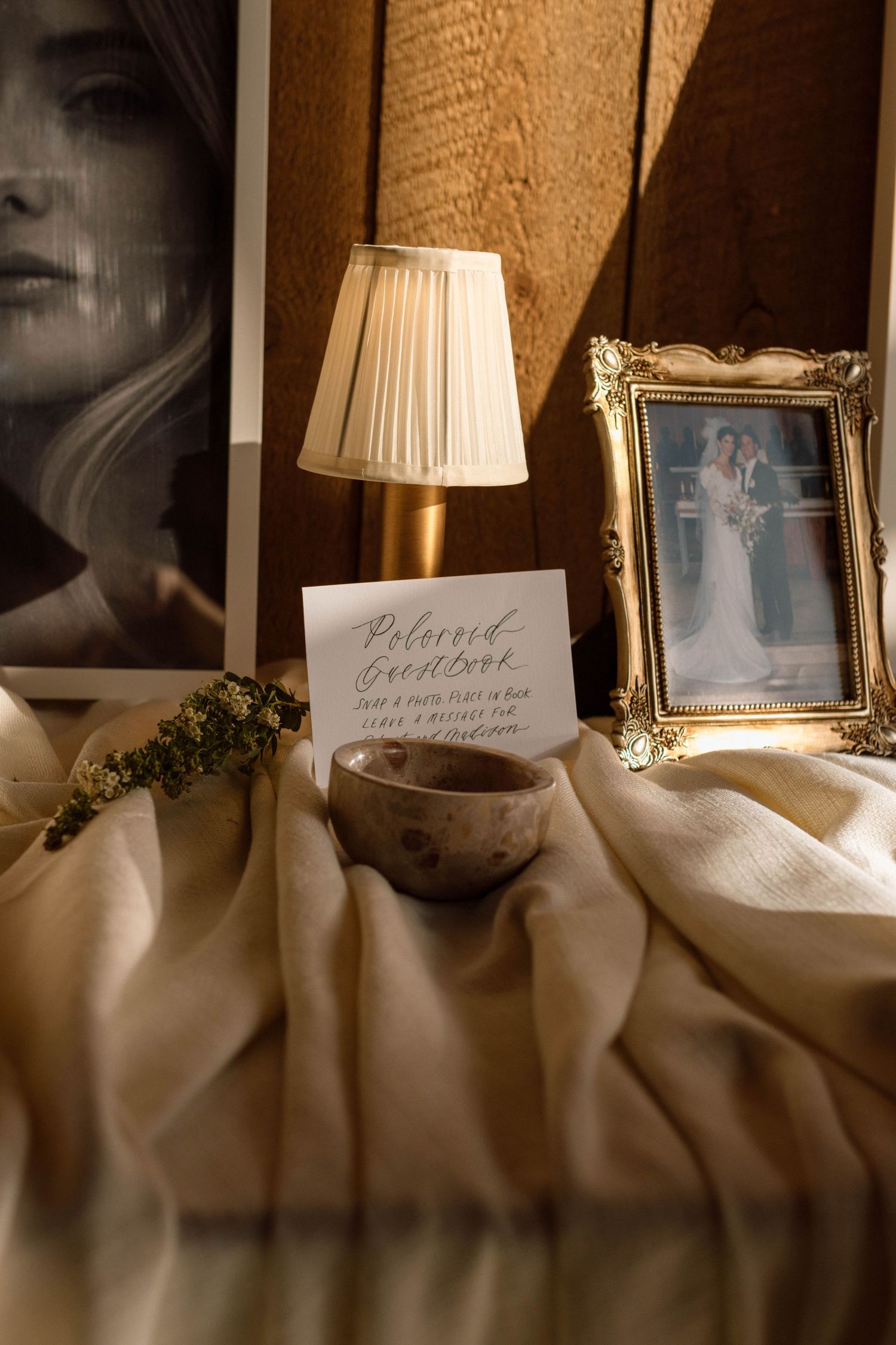 Elegant wedding guest book display with a small lamp, calligraphy sign, ceramic bowl, and gold framed photo on draped linen.