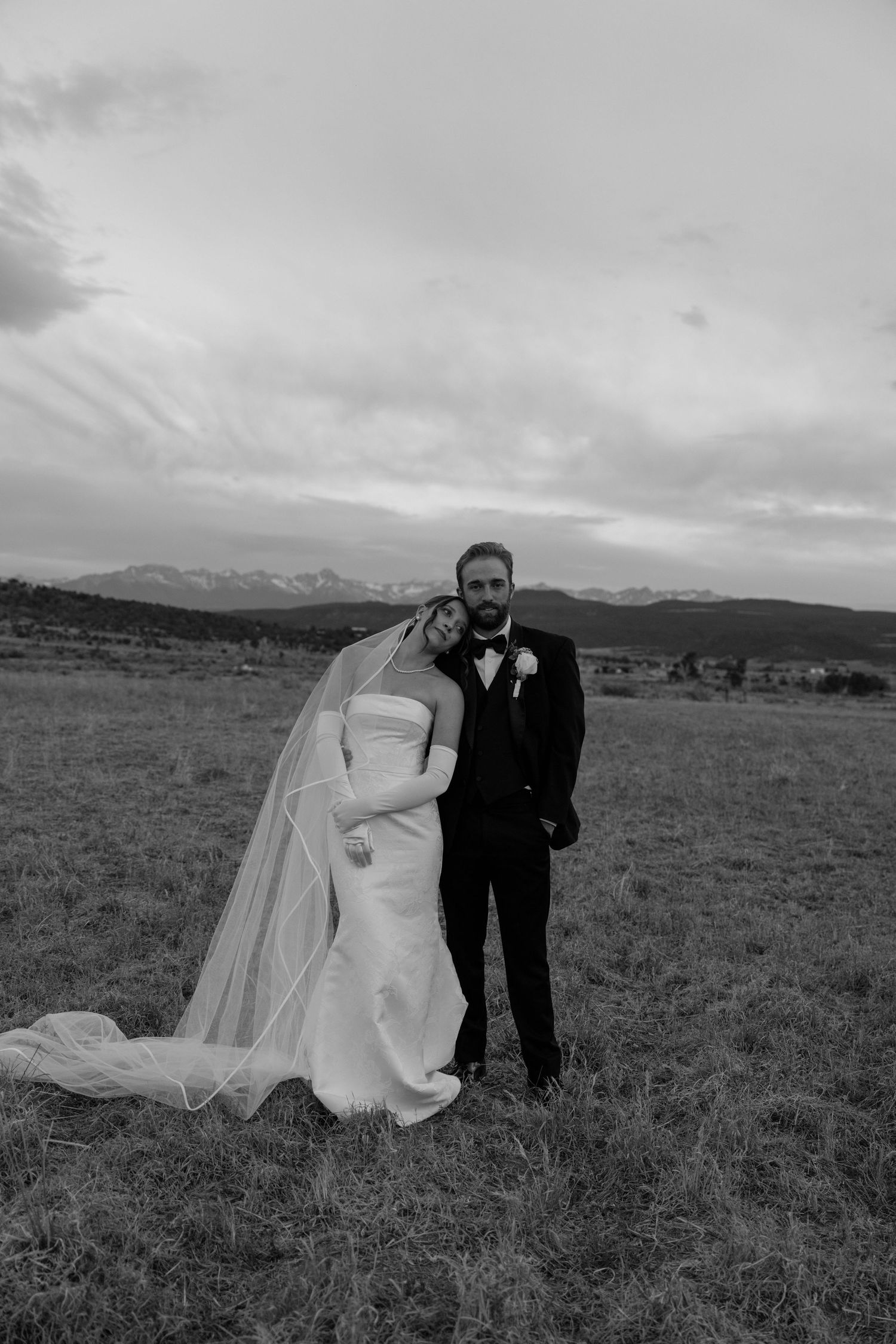 Black and white photo of a bride and groom posing in a mountain meadow, bride in strapless gown with veil, groom in tuxedo.