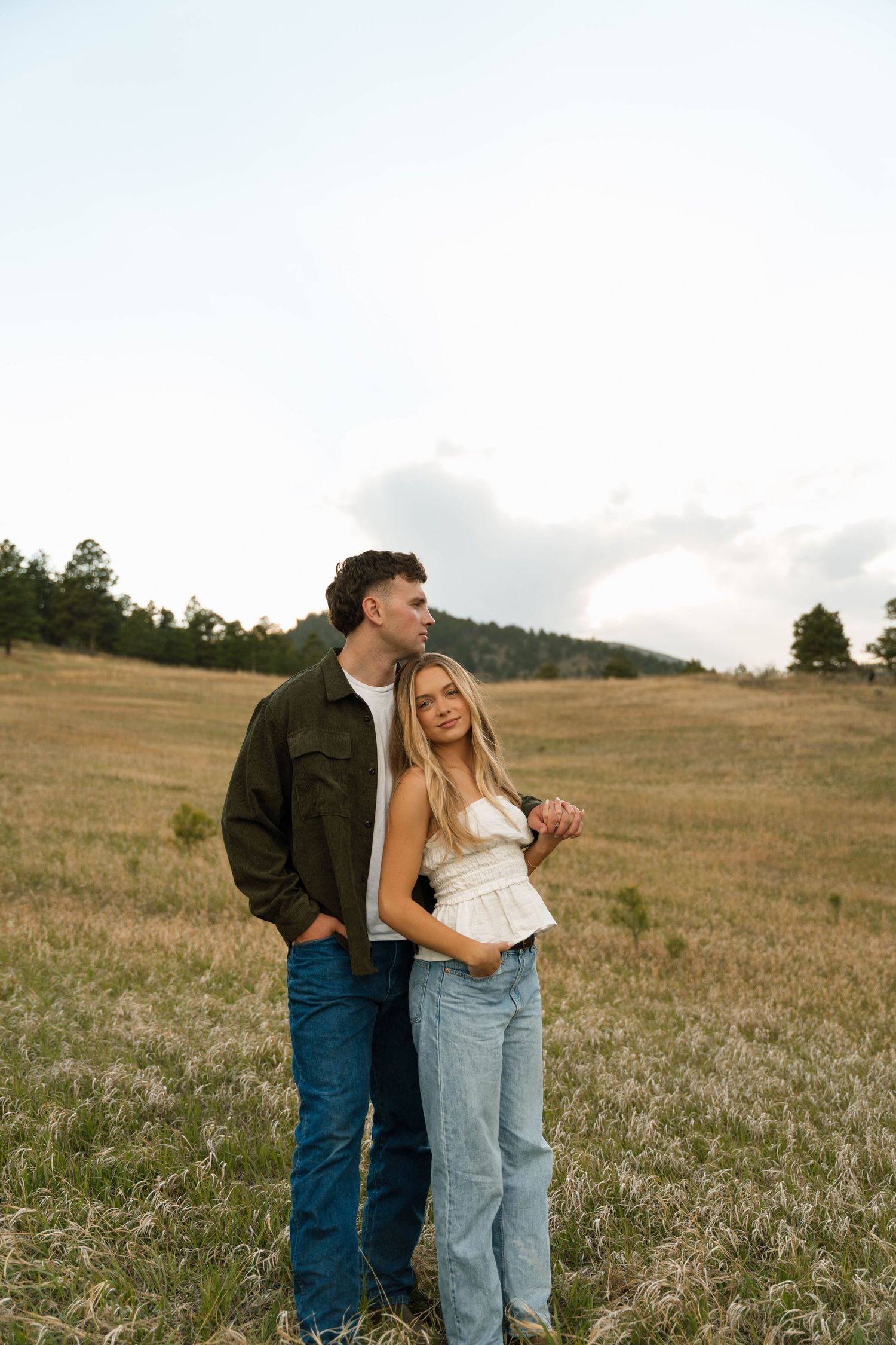 A couple embraces in a scenic meadow, man in green jacket kissing woman's head, both wearing jeans.
