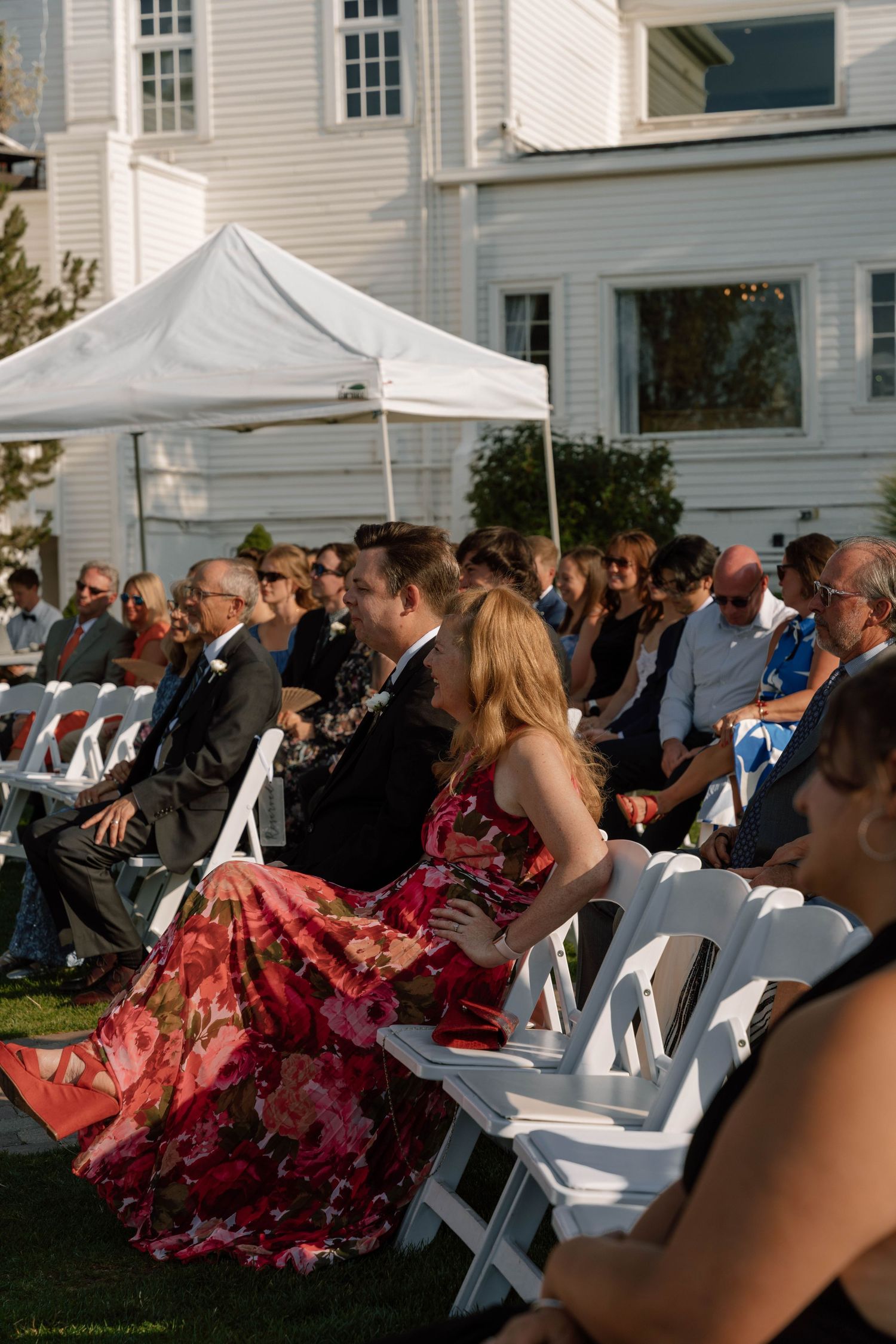 Wedding guests seated on white chairs on a lawn beside a historic white house.