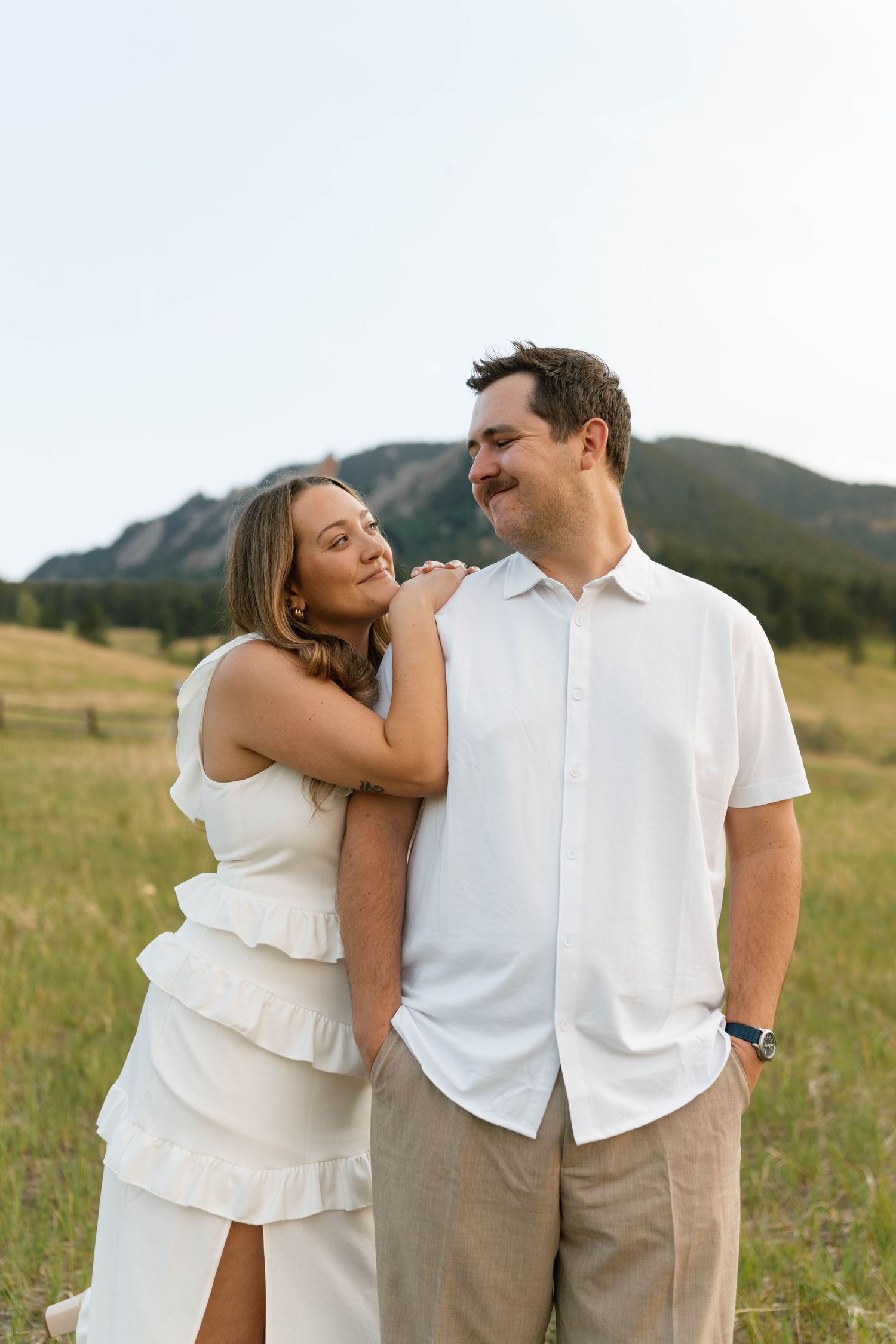 Woman in white dress leaning on mans shoulder in sunny mountain meadow with Flatirons in background.