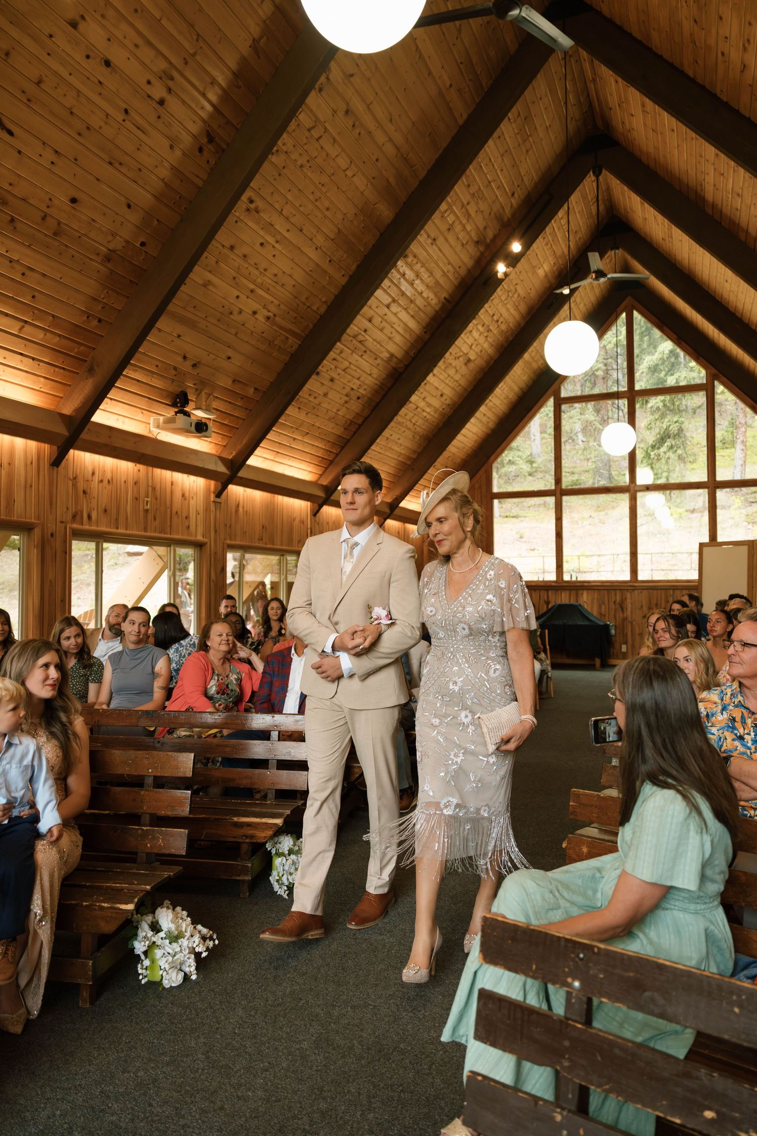Groom escorts mother down the aisle of a wood-paneled chapel filled with wedding guests.