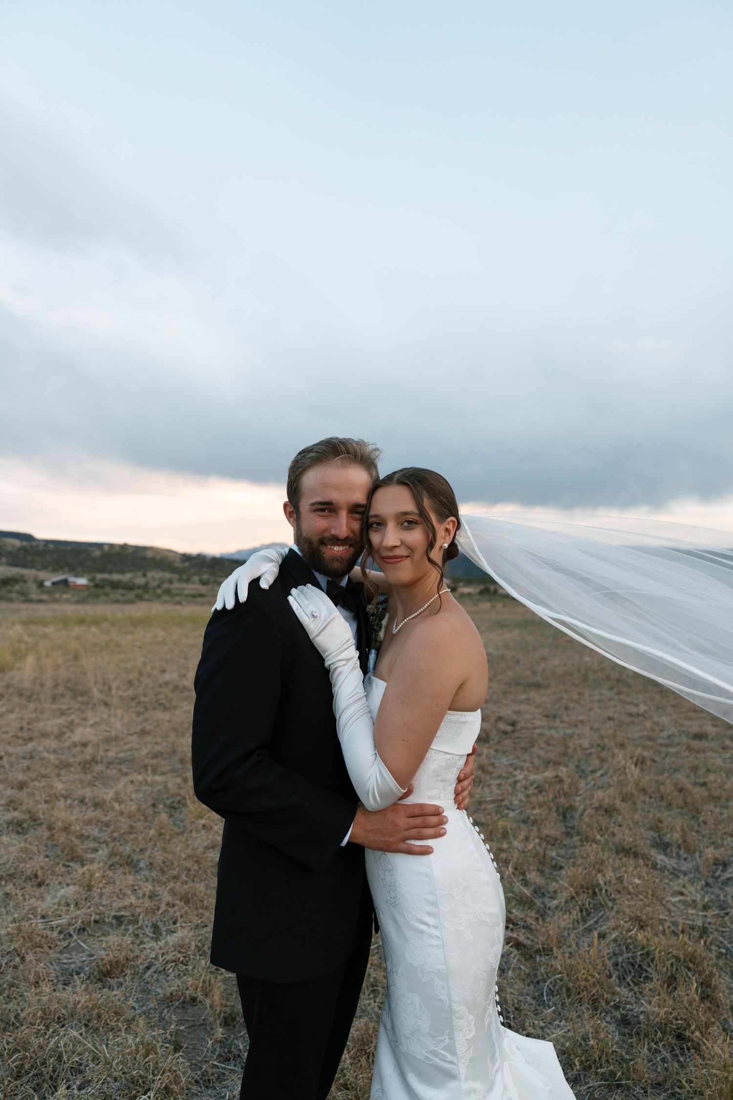 Bride in white gown and veil embraces groom in black tuxedo outdoors at dusk in an open field.