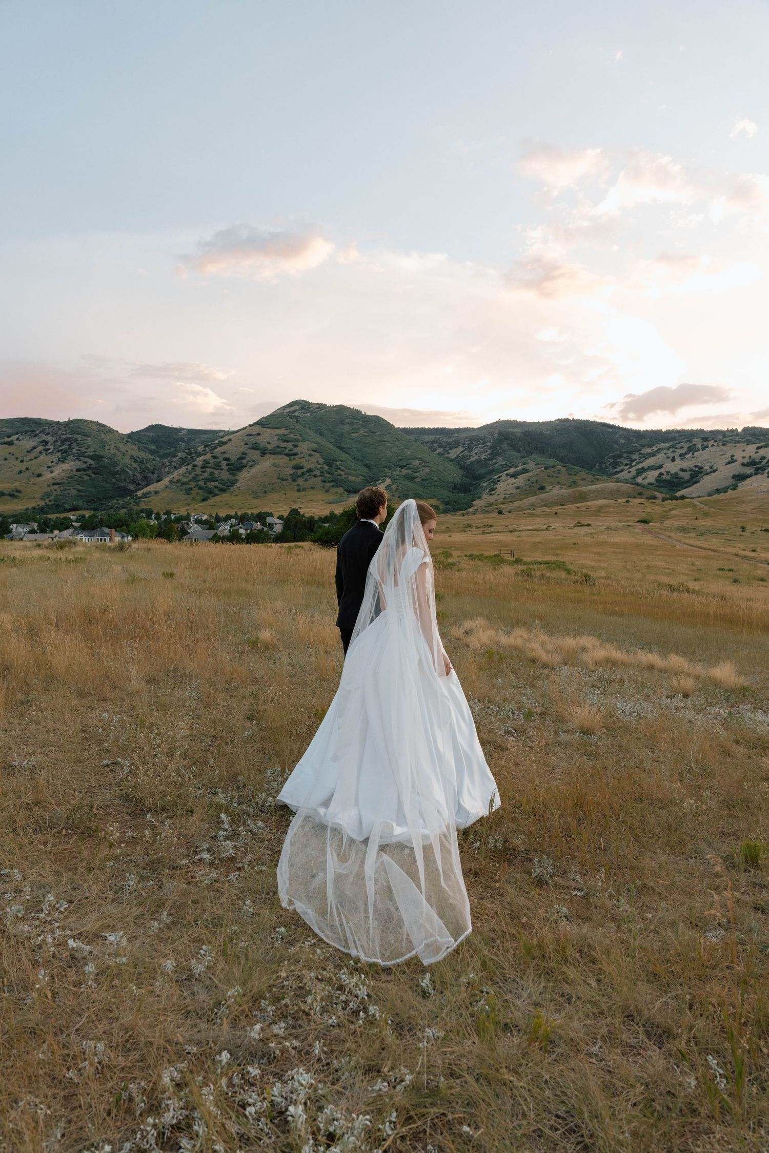 Long flowing wedding veil trails behind couple in mountain meadow during golden hour.