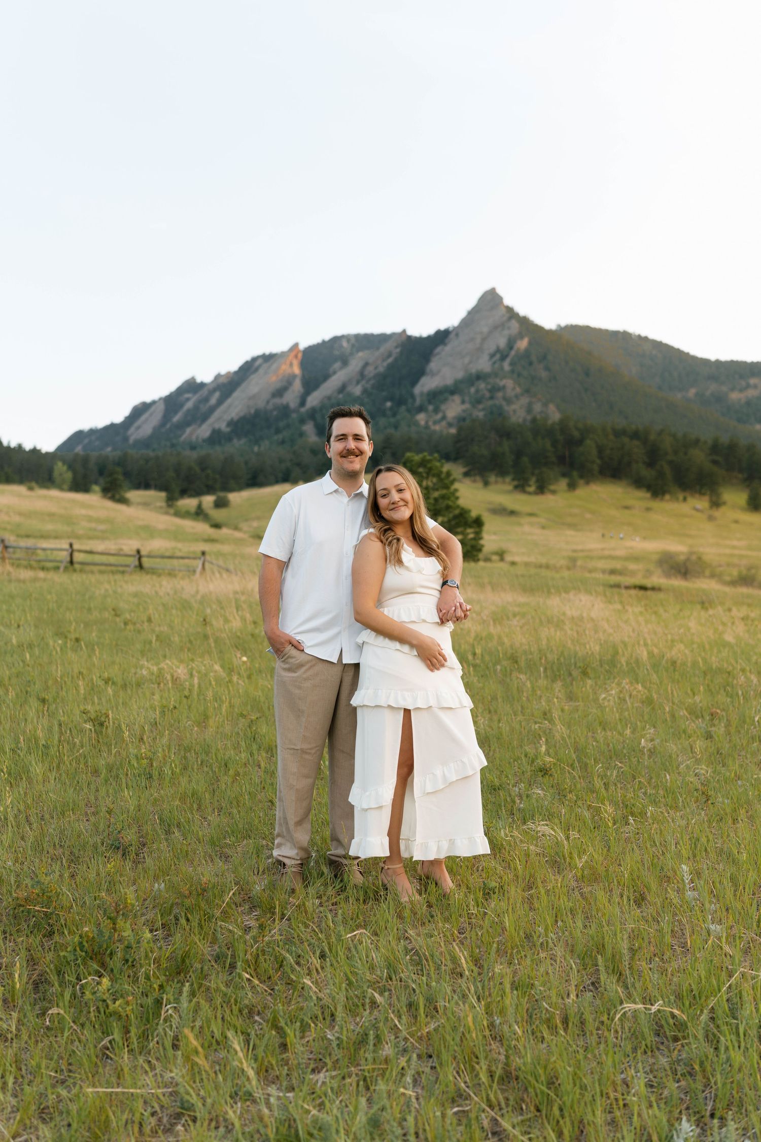 Smiling couple posing together in a sunlit meadow with the Flatirons rock formation visible in the background.