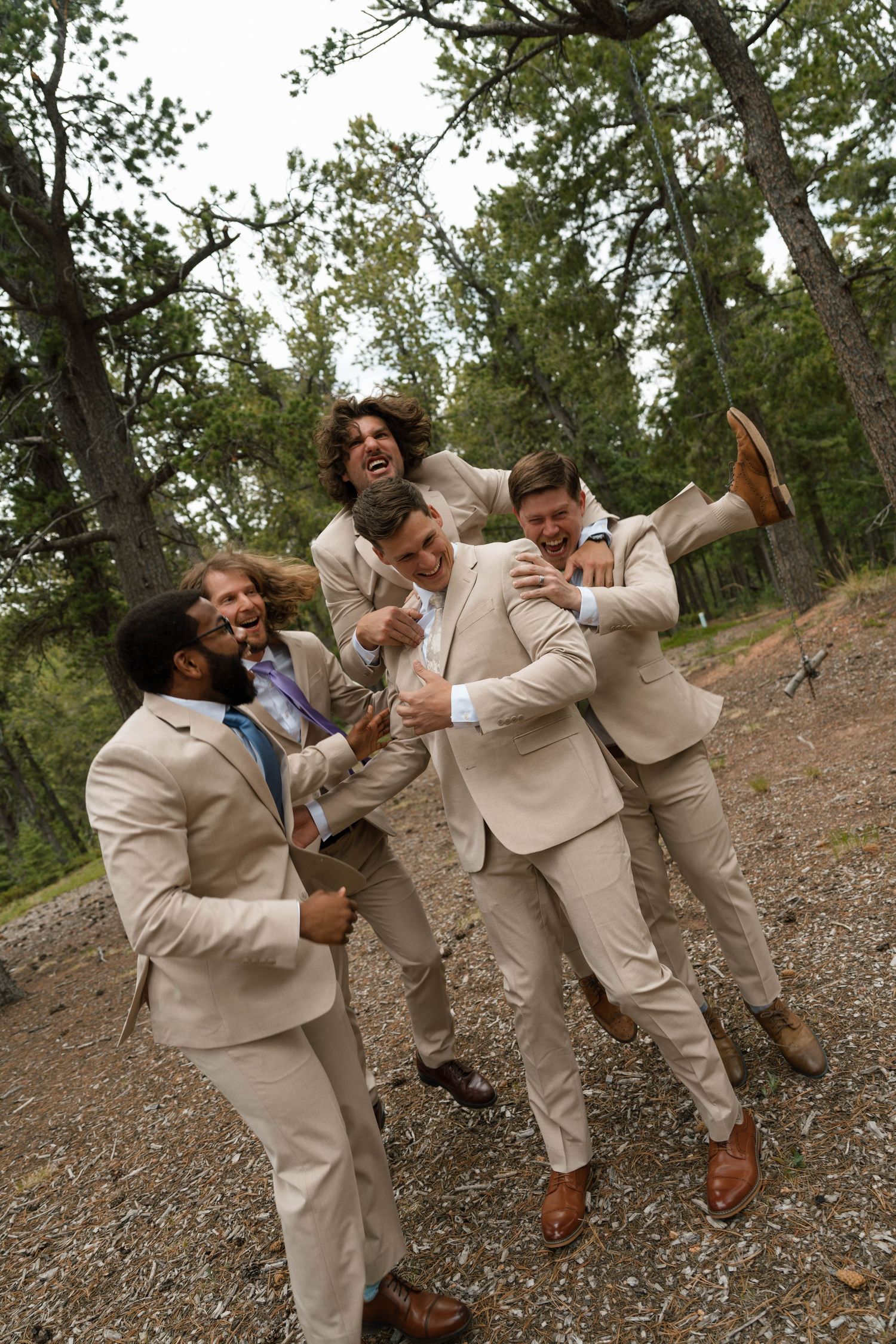 Four groomsmen in tan suits laughing and celebrating together in a forest setting.