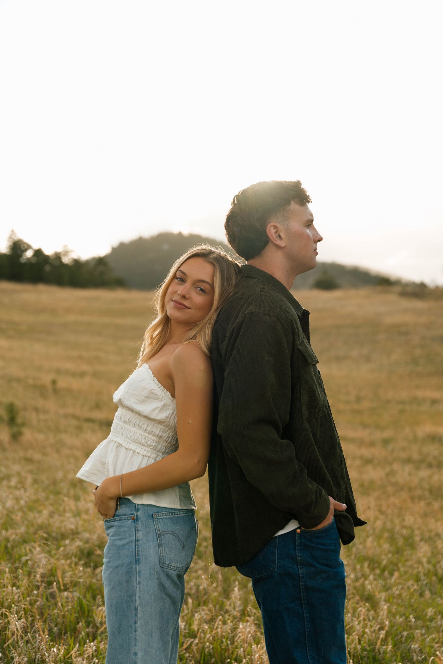 A couple stands back-to-back in a golden field at sunset, woman in white top and jeans smiling at camera.