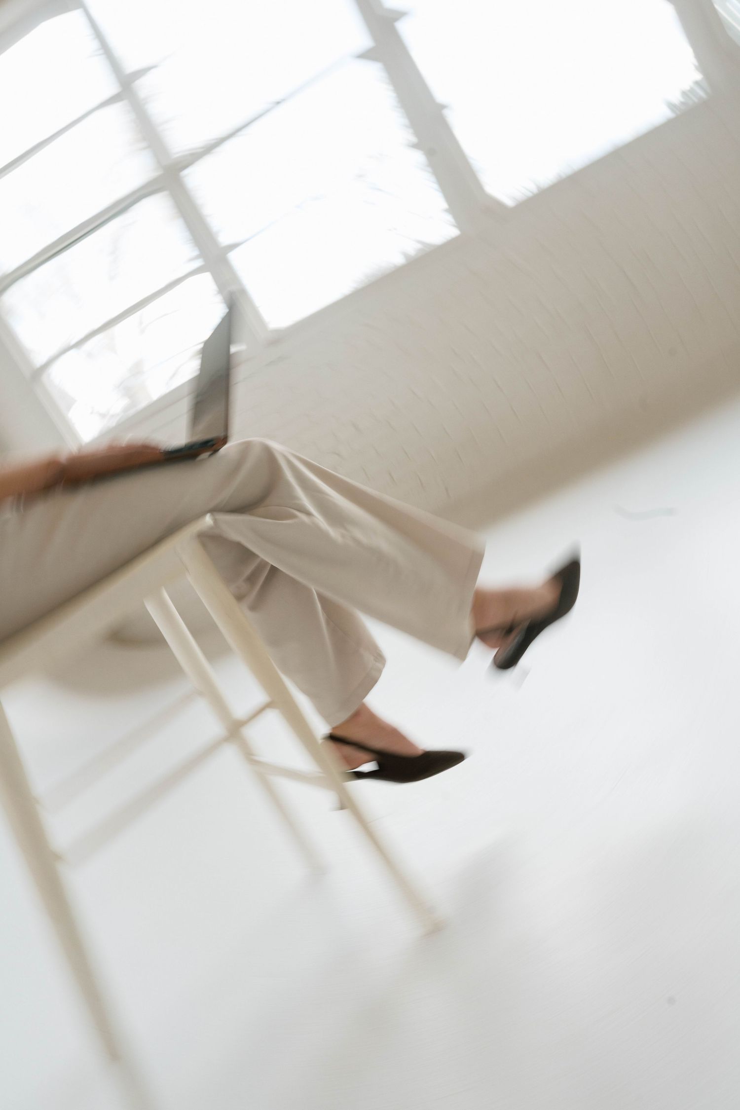 Blurred motion shot of person in white wide-leg trousers and black flats seated on a wooden stool in a bright white room.
