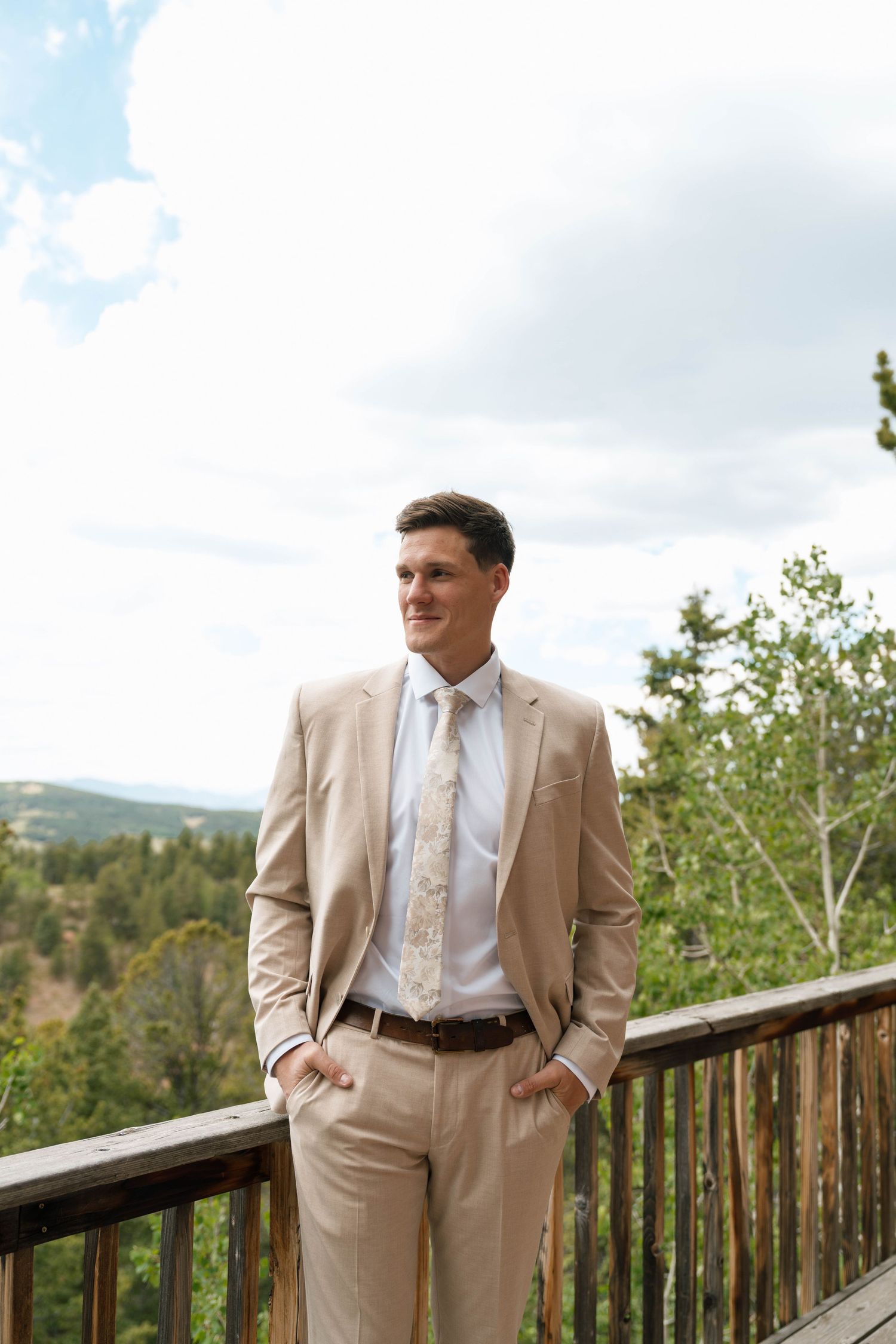 Groom in beige suit and floral tie stands on wooden deck with mountain forest backdrop.