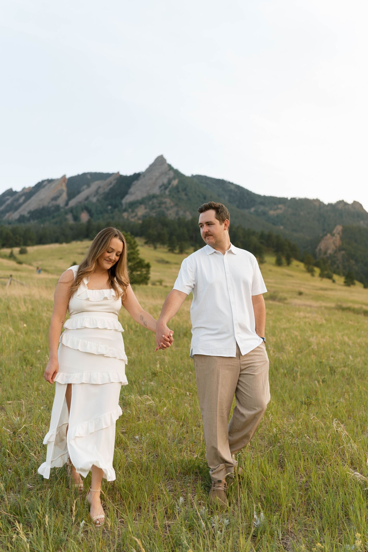Couple holding hands walking through grassy field with rocky Flatirons mountains in background, Boulder CO.