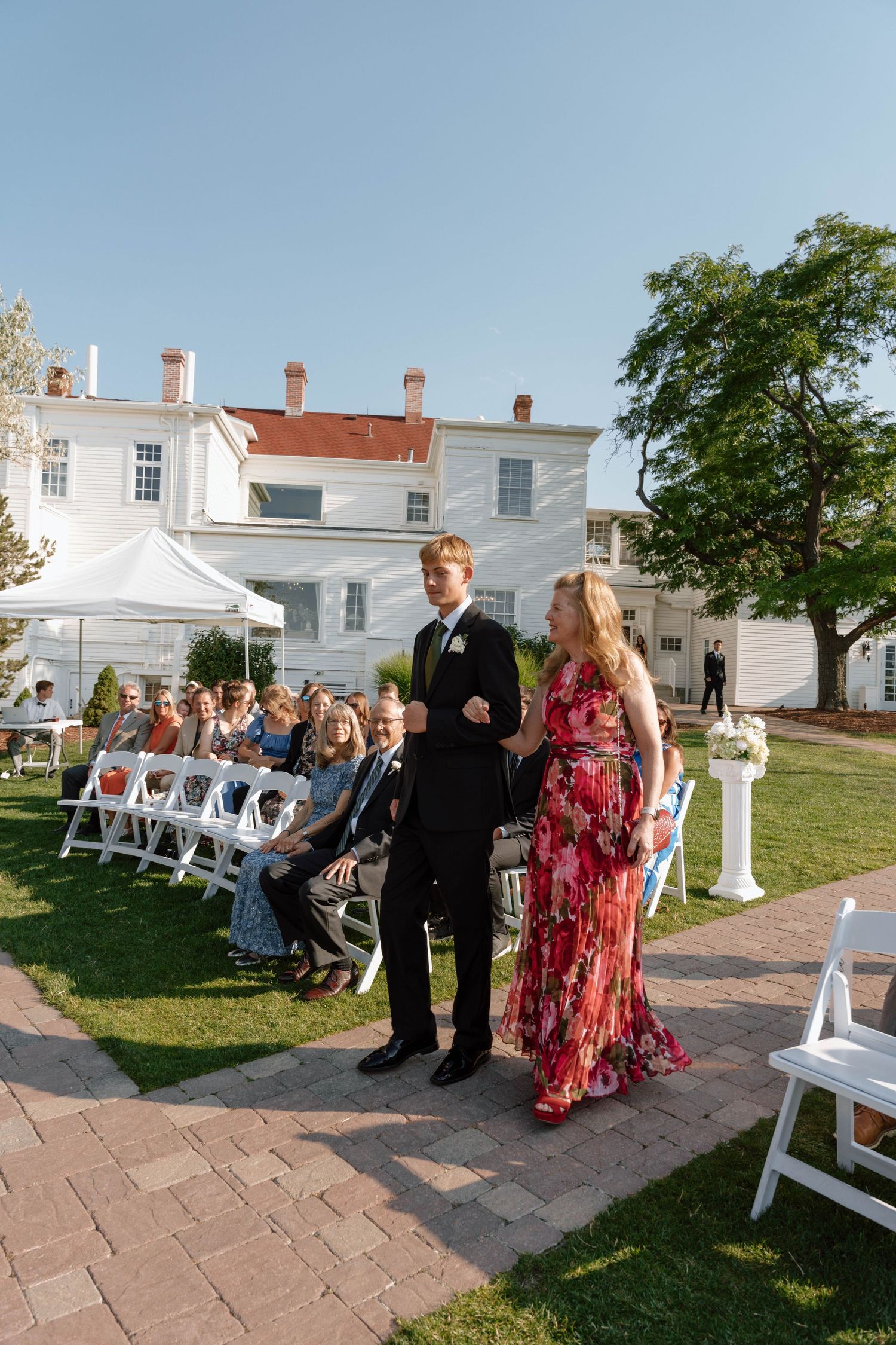 An outdoor wedding ceremony with guests seated on white chairs and a historic white house in the background.