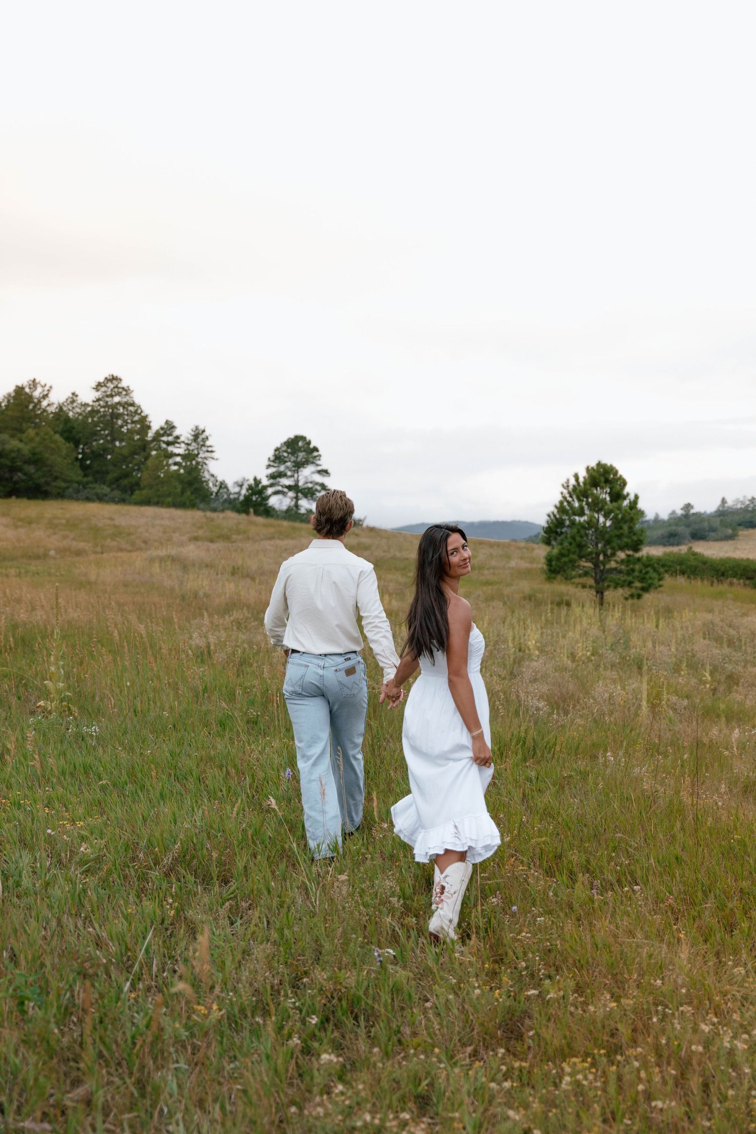 A couple walks hand in hand through a scenic meadow wearing white and denim on an overcast day.