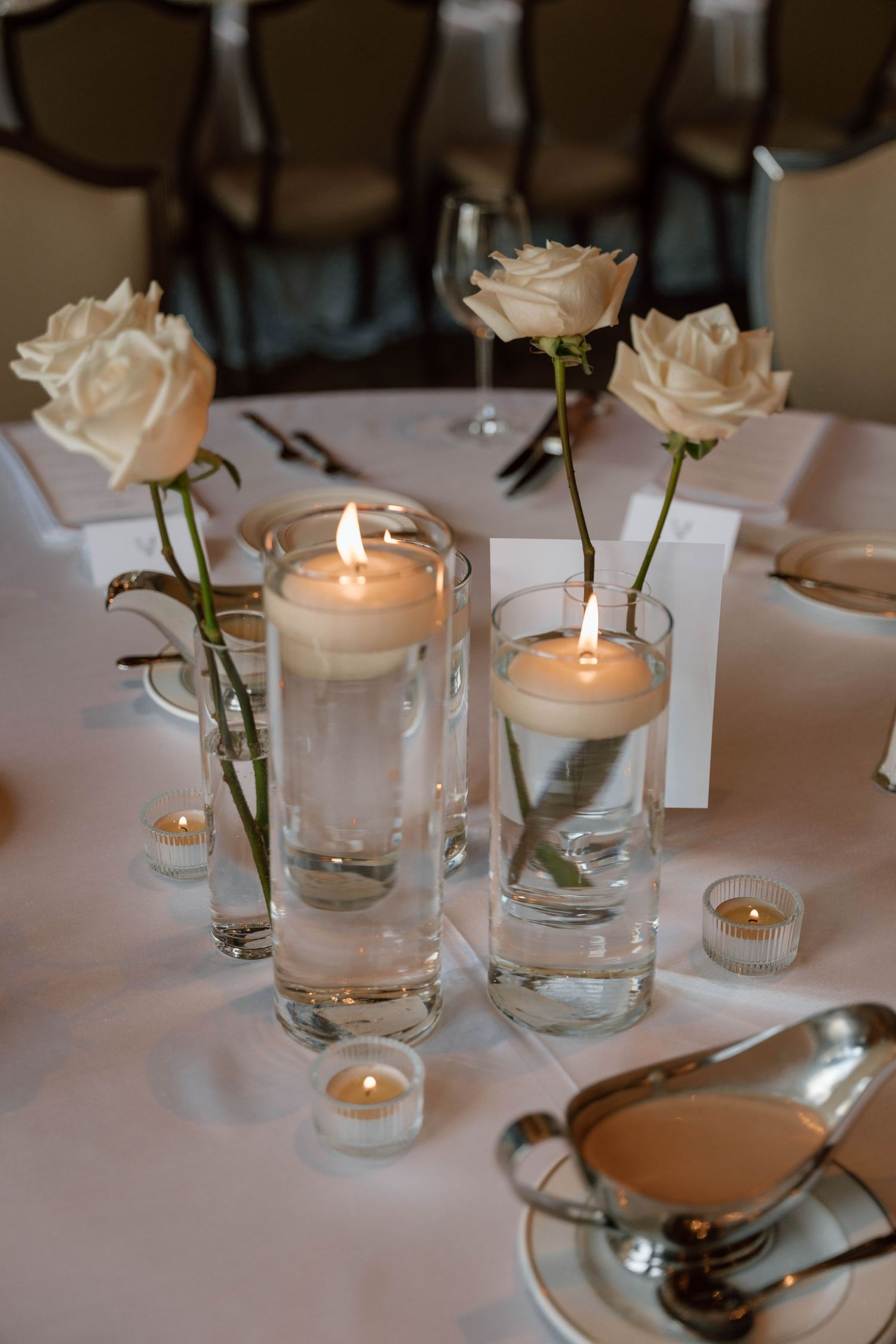 Elegant table settings with white roses and glowing candles in glass holders on white tablecloths.