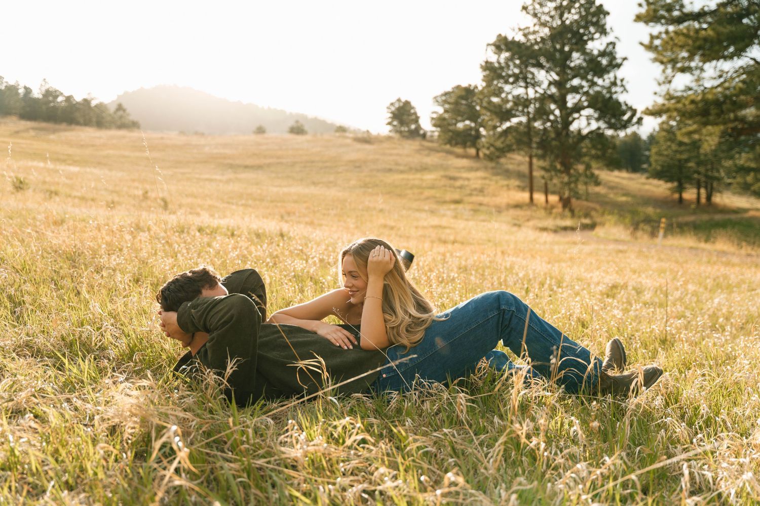 Couple lying together in a sunny golden meadow with rolling hills and pine trees in the background.