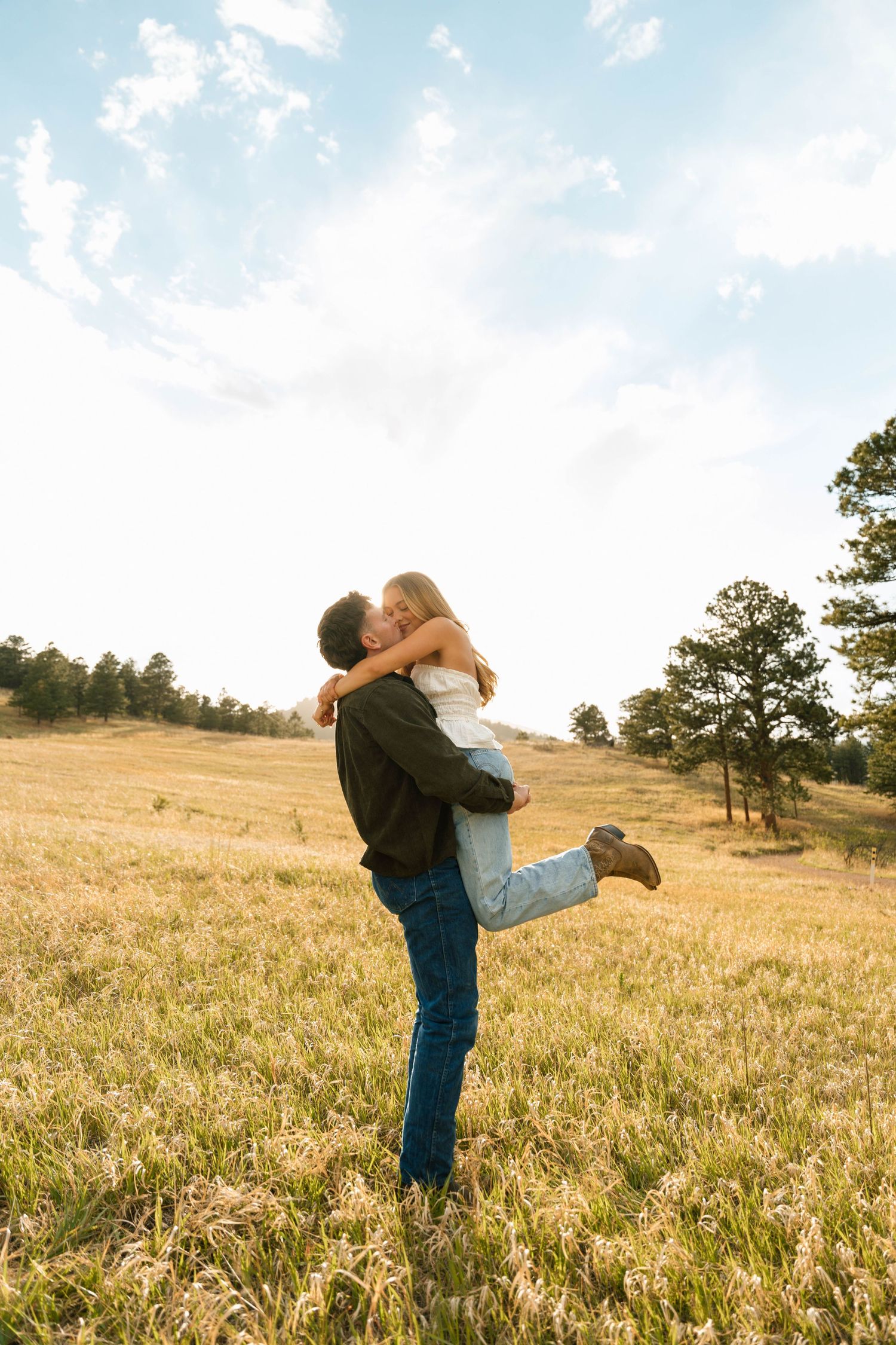 A couple shares a romantic embrace in a golden meadow, the man lifting the woman as they kiss under a partly cloudy sky.