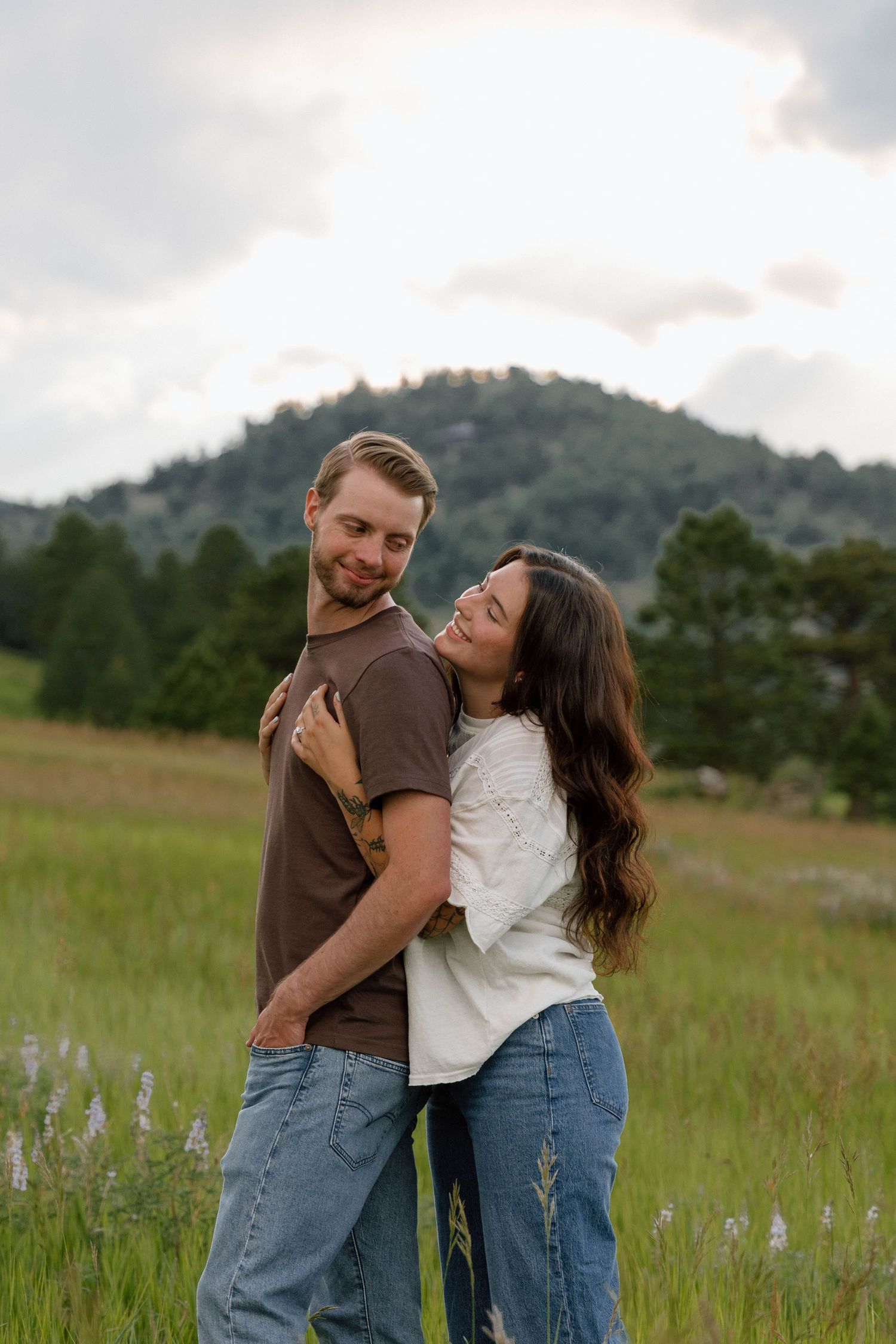 Couple sharing laughter and embraces in a sunlit field with rolling hills behind them.