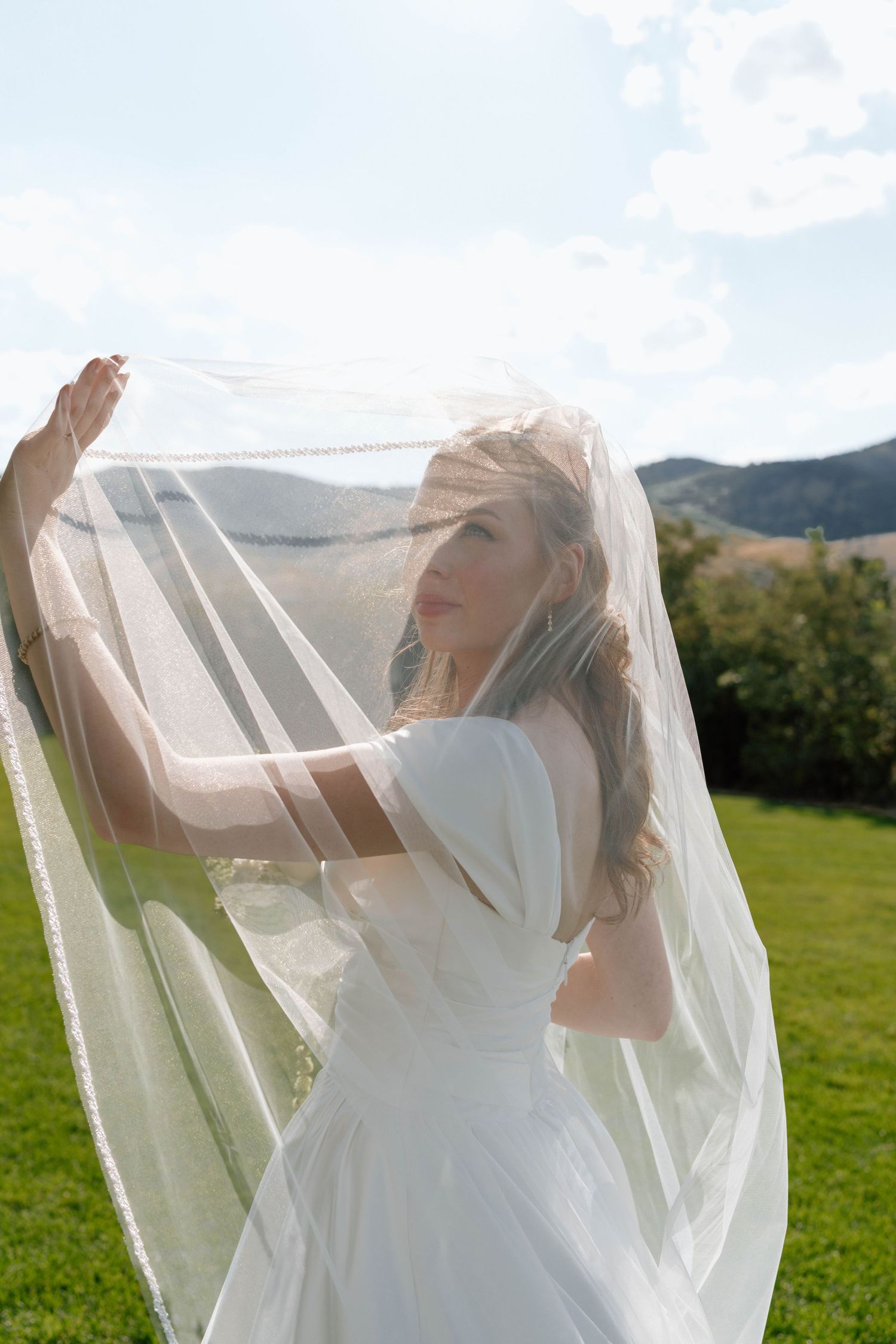A bride in white dress plays with her veil in a sunny meadow with mountains in background.