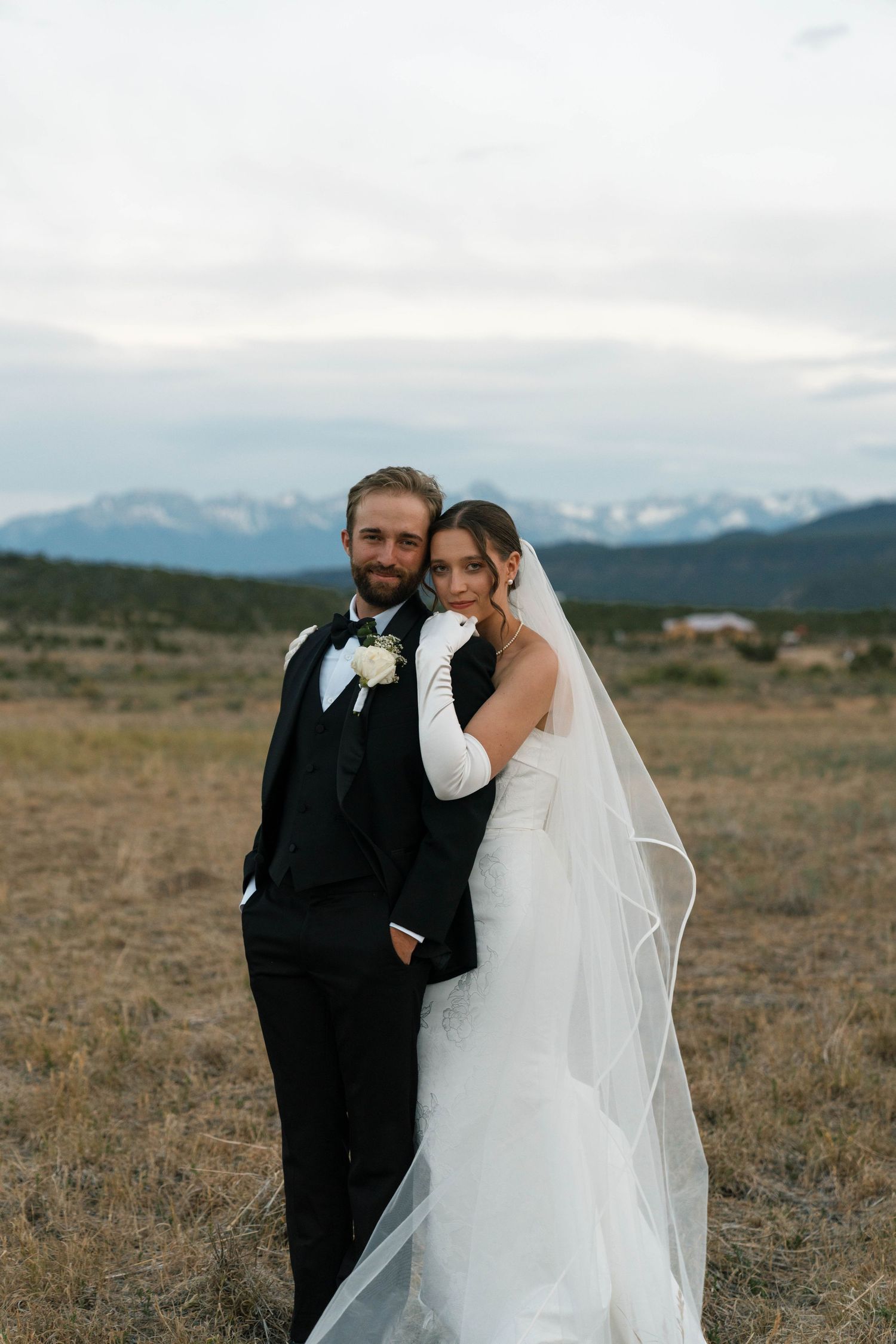 Bride and groom pose romantically in open field with snow-capped mountain backdrop at dusk.