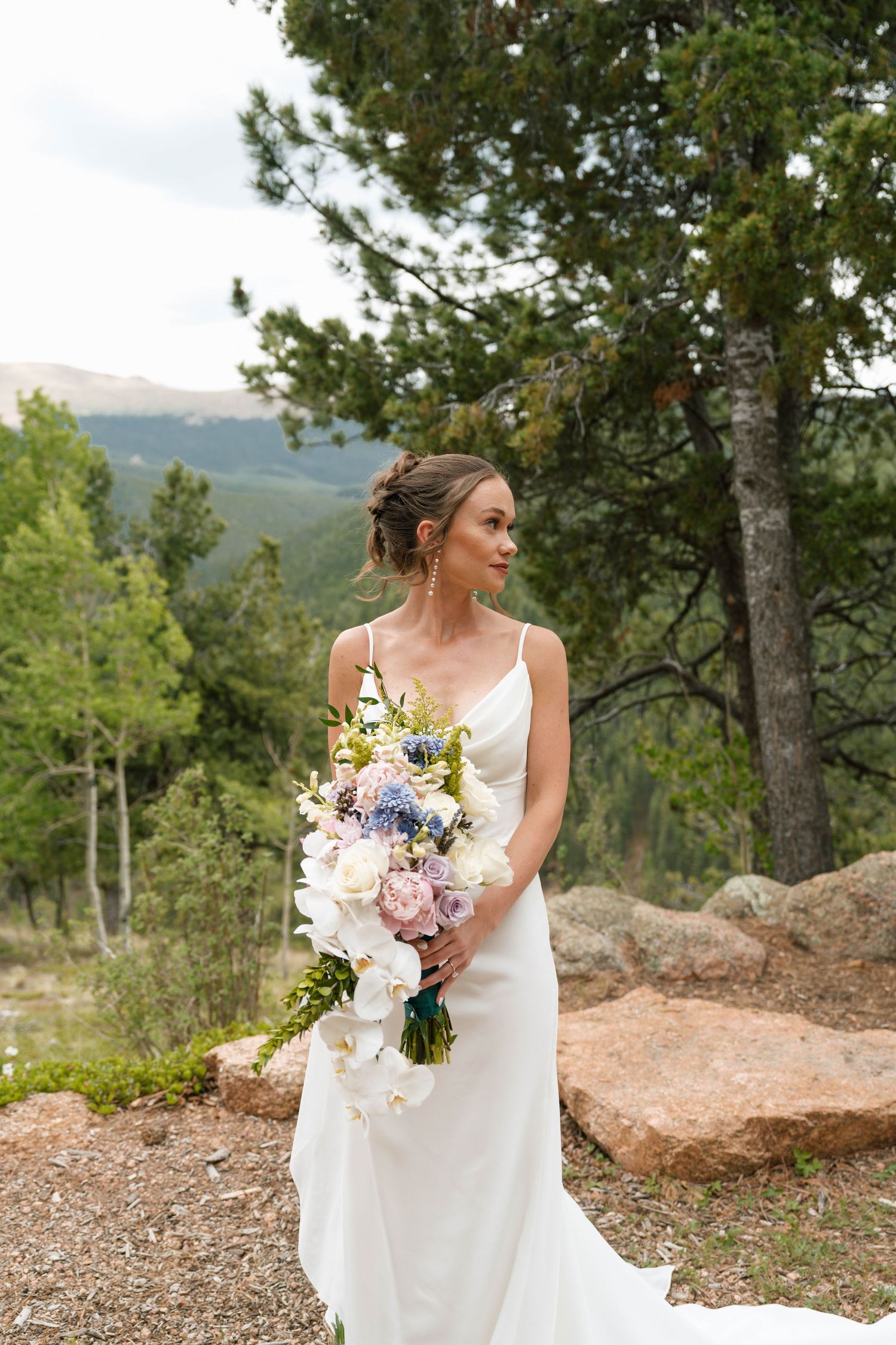Bride in white gown holding colorful bouquet with mountain forest backdrop.