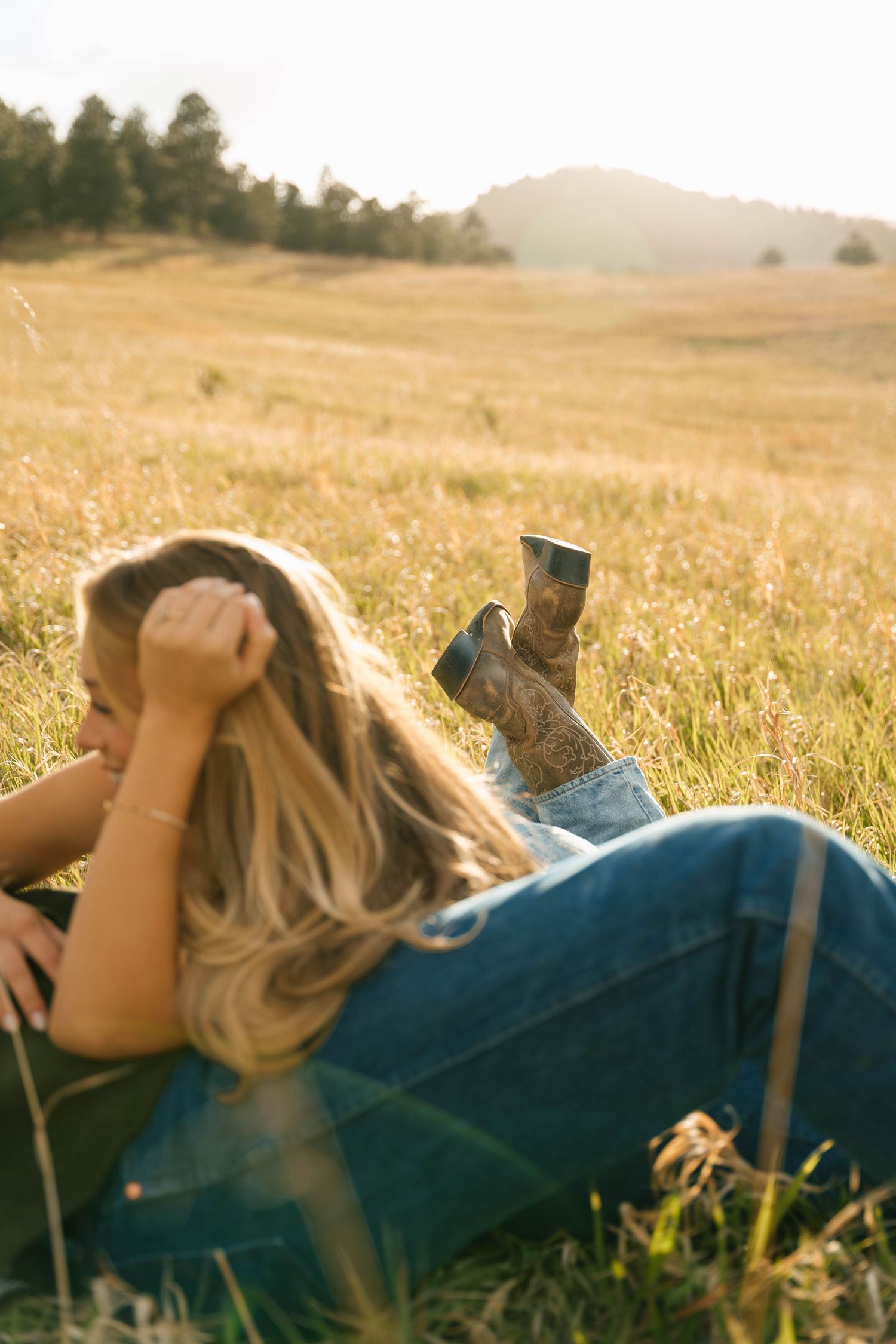 Woman with blonde hair lying in golden grass field wearing jeans and cowboy boots, bathed in warm sunset light.