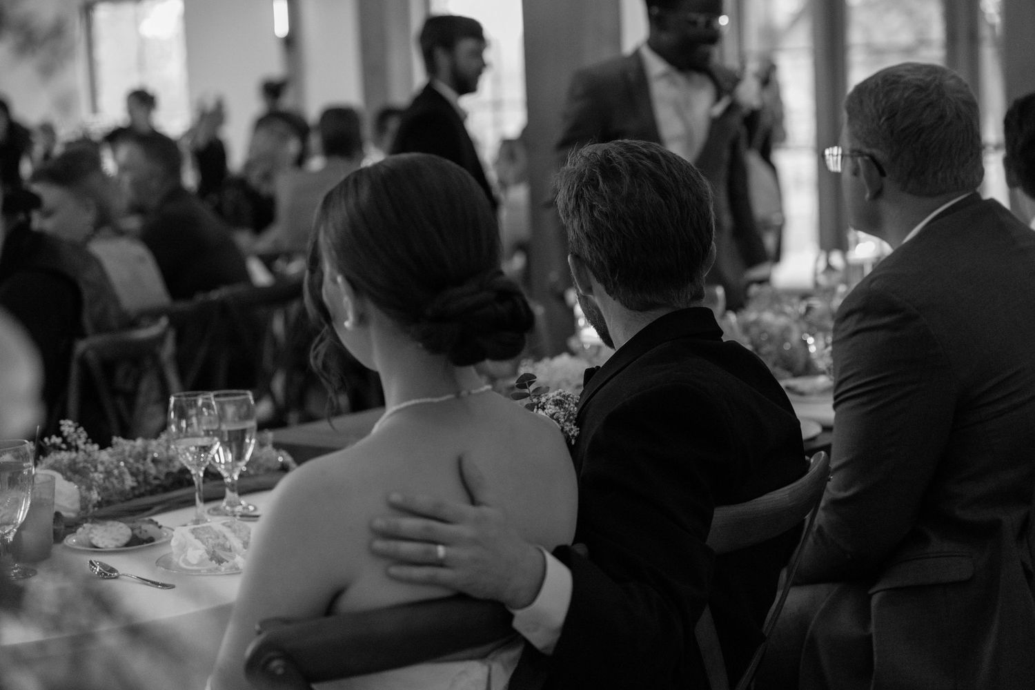 Black and white photo of wedding guests seated at a reception table, with a man resting his hand on a womans bare shoulder.
