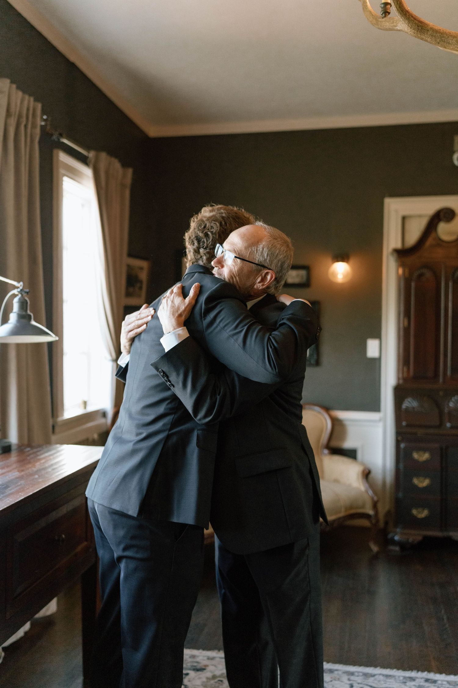 Two people in formal attire share an emotional embrace in an elegantly furnished room.