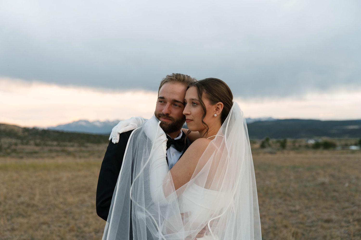 Bride and groom embracing in a golden field with mountain backdrop at sunset on their wedding day.