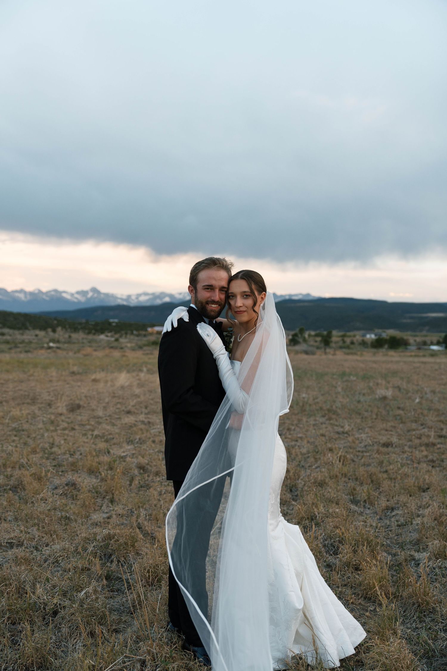 Bride and groom embracing in golden field with snow-capped mountains at dusk.