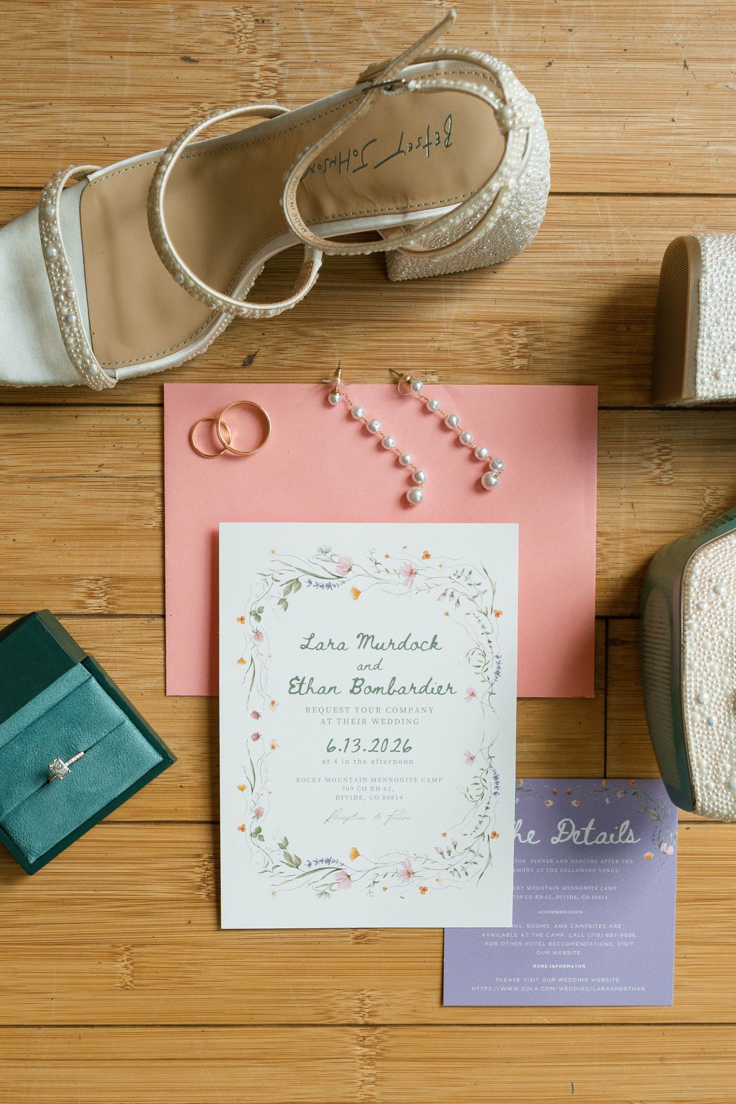 Wedding flat lay with floral invitation, gold rings, pearl earrings, bridal heels, and teal ring box on wood floor.