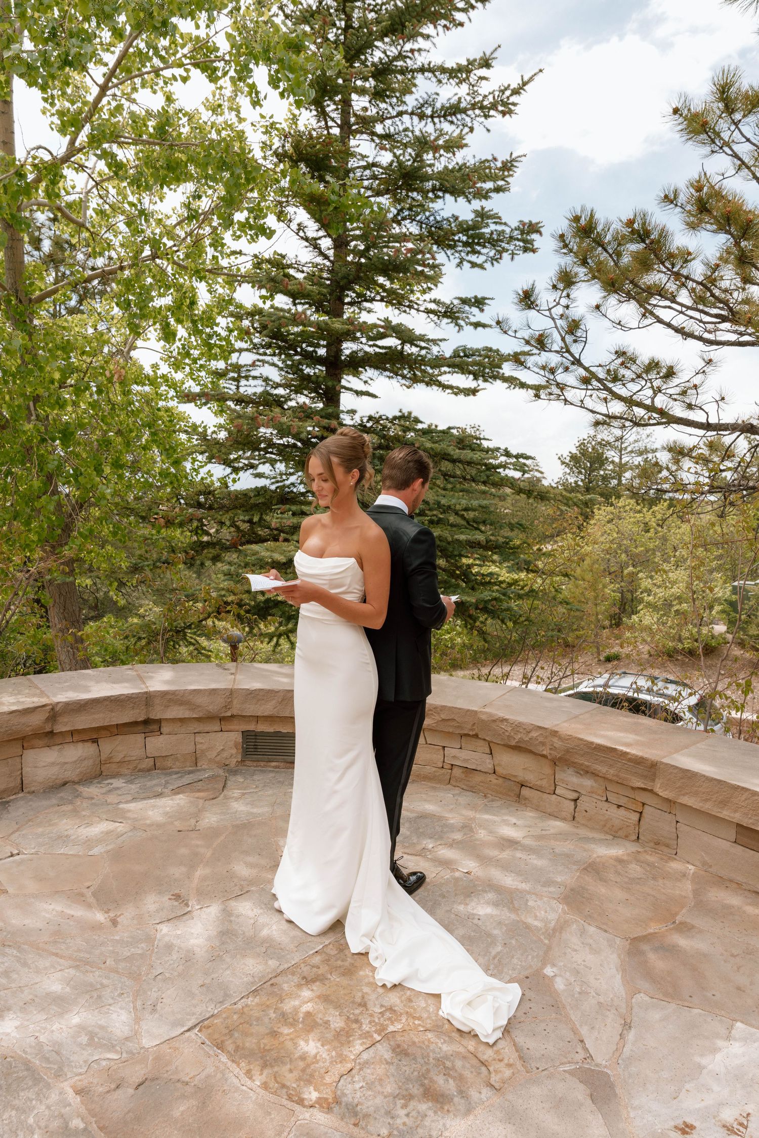 Couple in formal attire standing back-to-back on stone terrace surrounded by pine trees.